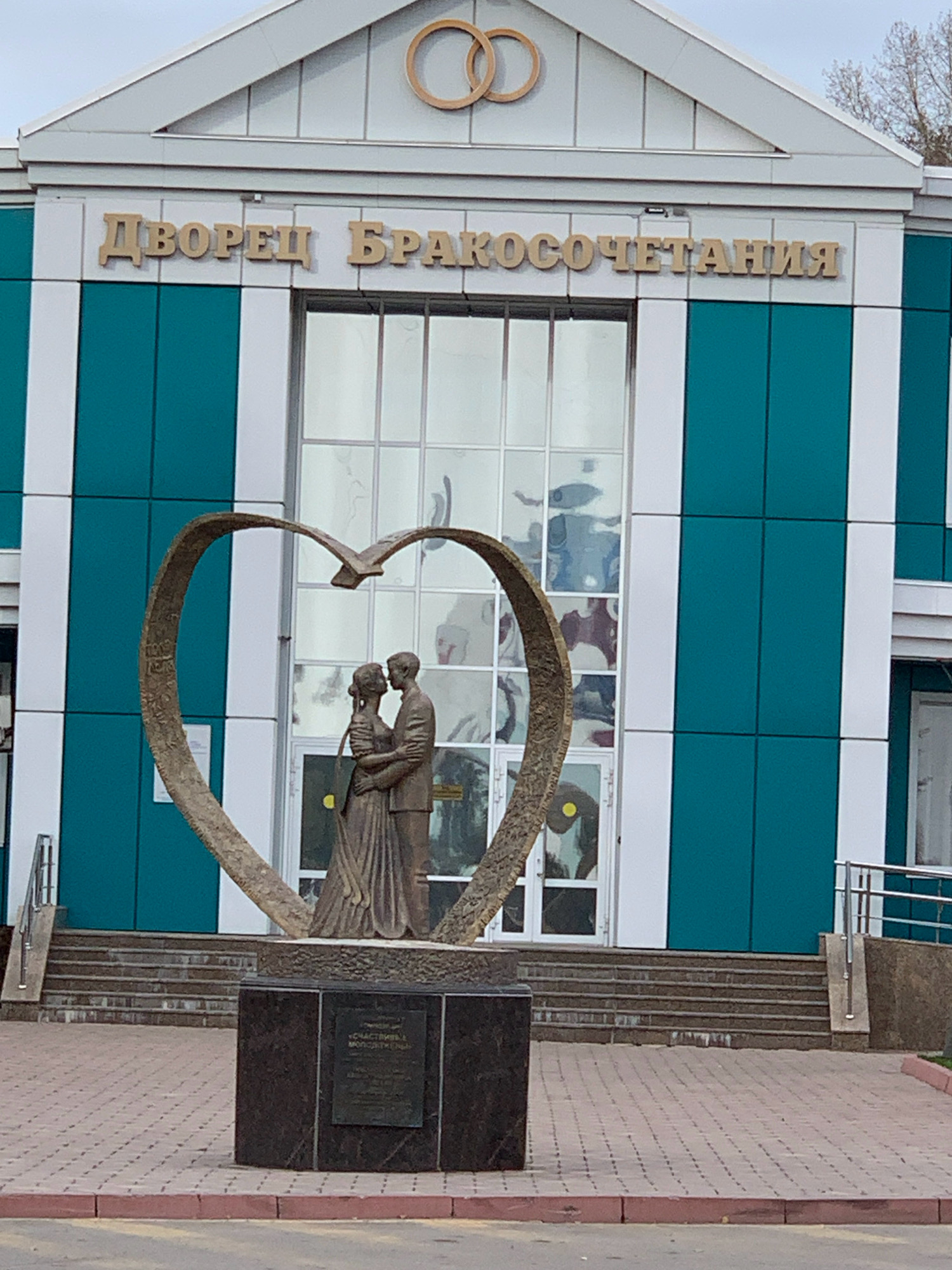 The image shows a sculpture of a couple kissing within a heart-shaped arch in front of a building with a sign that reads "ДВОРЕЦ БРАКОСОЧЕТАНИЯ" (Palace of Weddings) in gold lettering. The building has a modern facade with teal and white panels and large glass windows. The sculpture is made of bronze and stands on a dark granite pedestal. Behind the sculpture, steps lead up to the entrance of the building. The area in front of the building is paved with brick. This is the Palace of Weddings in Prokopyevsk, Russia.