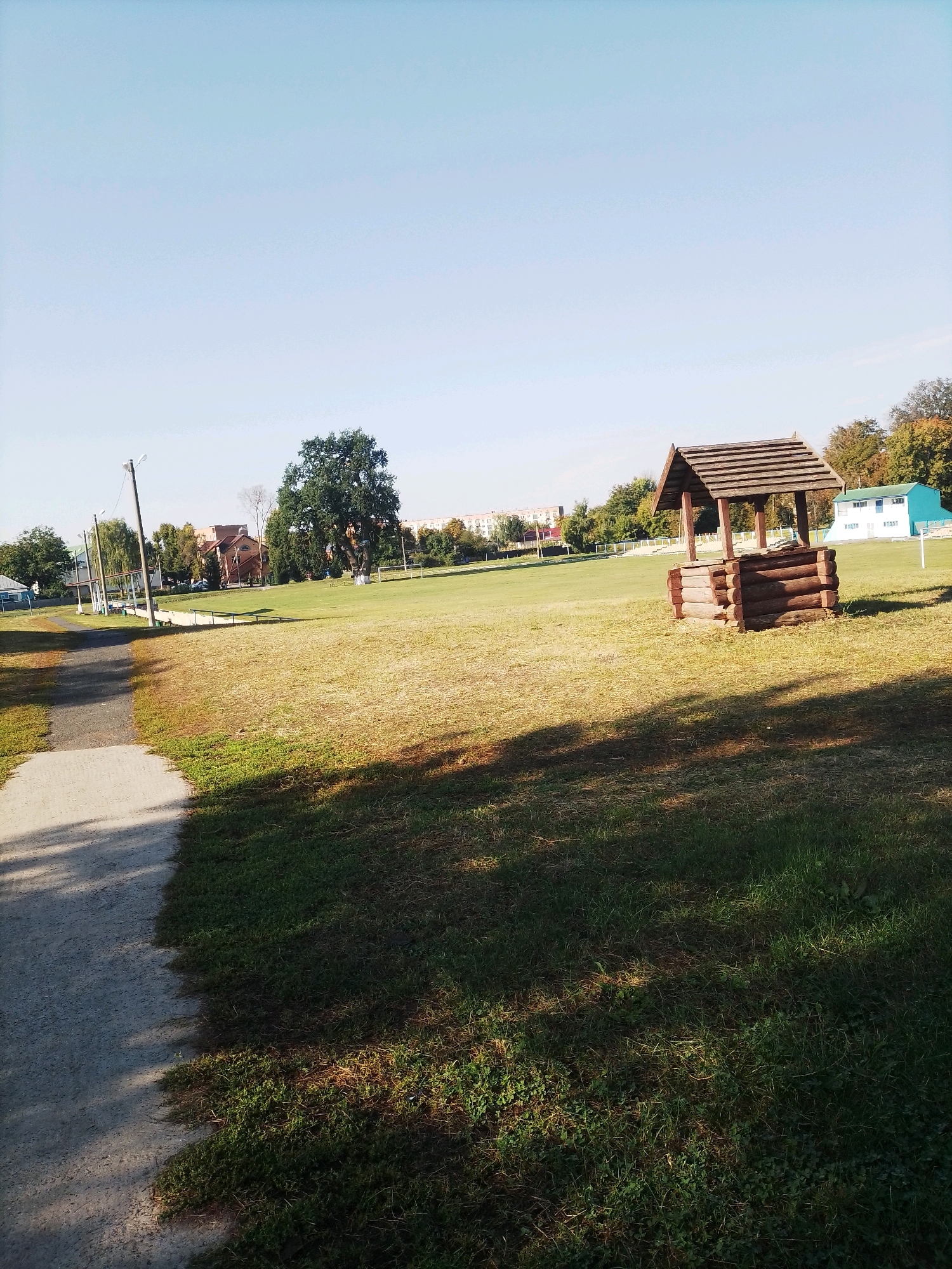 The image depicts an outdoor scene in Velyka Bahachka, Ukraine. In the foreground, a paved pathway runs along the left side of the image, casting a shadow onto the grassy area beside it. The pathway leads into a large, open field with dry, yellowing grass. Dominating the mid-ground is a wooden well structure, constructed from logs and topped with a sloped, wooden roof. Further into the field, a large, mature tree stands prominently, its dense green foliage contrasting with the drier grass. In the background, various buildings are visible, including a low-rise brick building with a red roof, and a longer, light-blue building with white accents. The sky above is a clear, pale blue, indicating fair weather. The scene appears to be a park or recreational area, possibly near a school or sports facility, given the presence of a sports field marker in the far right background. No people are clearly visible in the image.