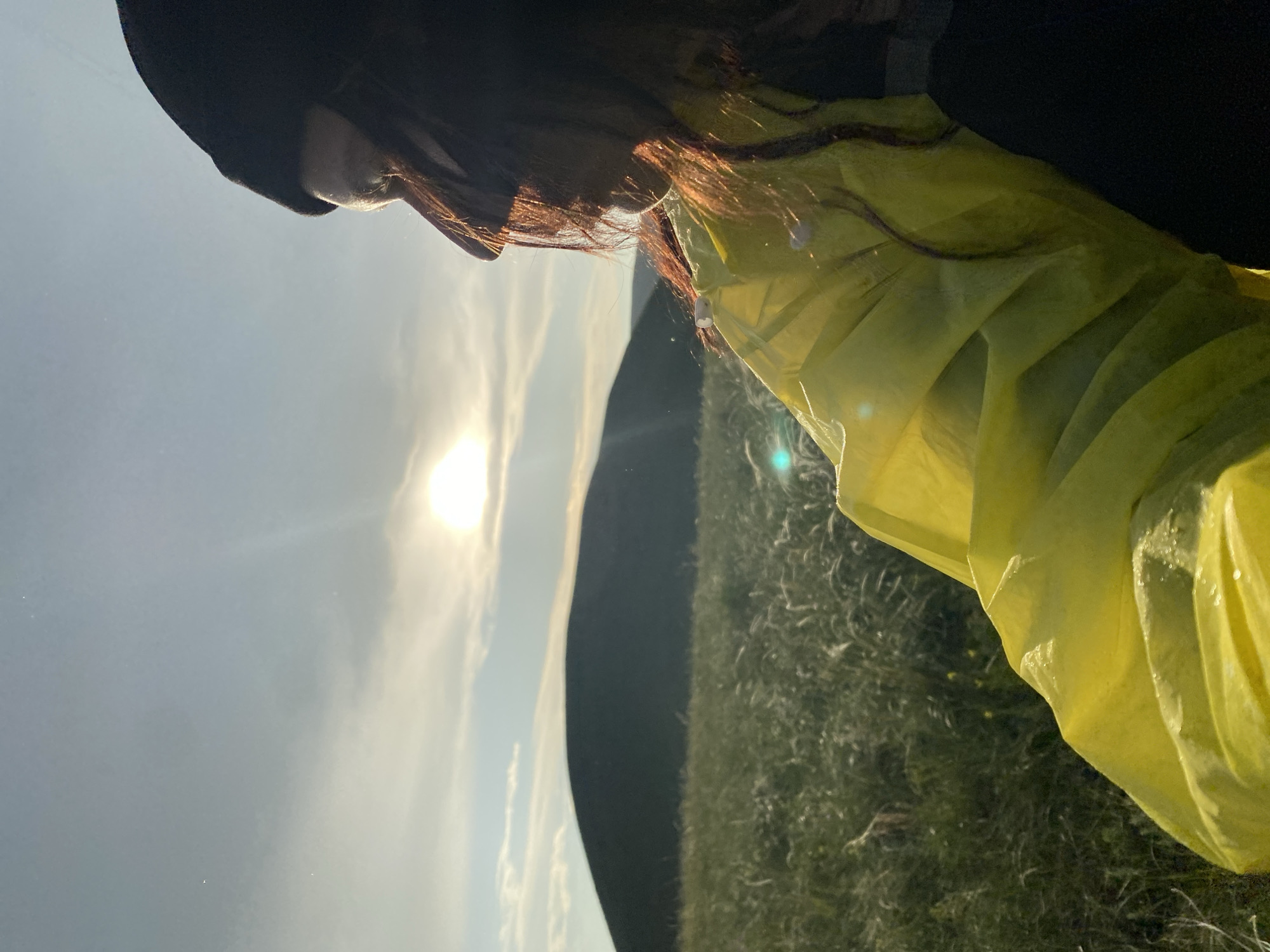 The image shows a person in a bright yellow jacket and black beanie, seen from the side and back, looking out at a natural landscape. The person's face is partially obscured by their hair and the angle of the shot, but they appear to be looking towards the right of the frame. The sun is visible in the sky, partially obscured by clouds and casting a warm glow. In the background, there is a dark, rounded hill or mountain, and a field of tall, wispy grass. The scene appears to be outdoors, possibly in a rural or natural setting. The location is specified as the city of Donskoye, Russia, suggesting this is likely a natural area within or near the city. The lighting and atmosphere suggest it might be late afternoon or early evening.