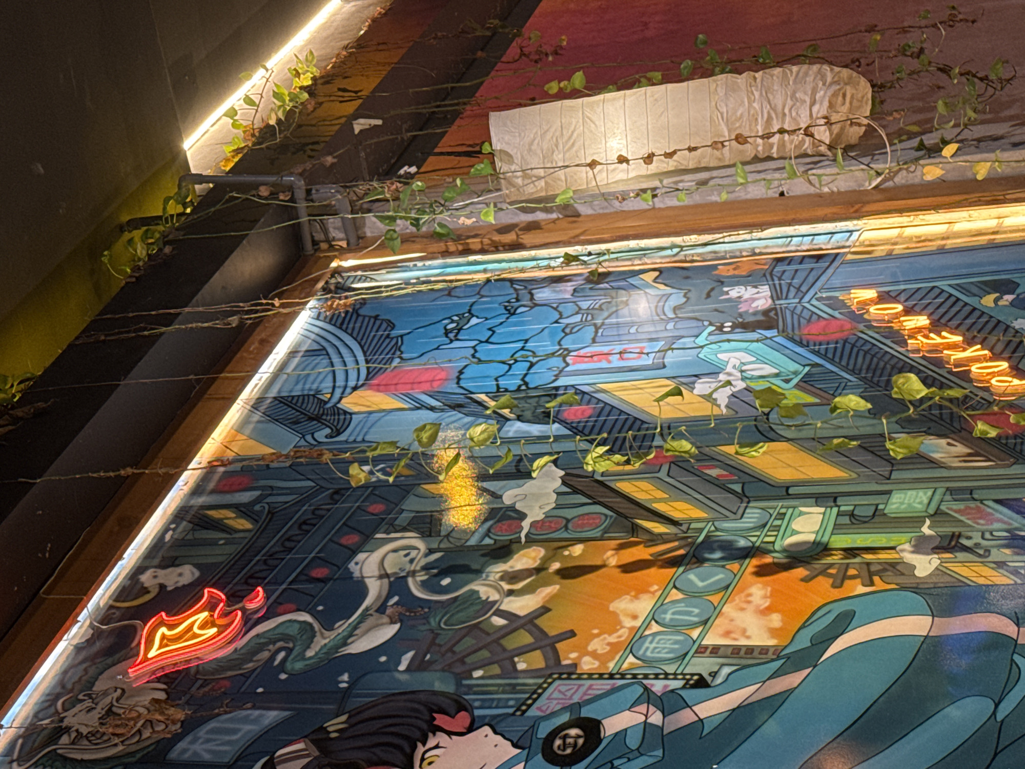 The image depicts a section of a wall in what appears to be a restaurant or bar, likely in Đà Nẵng, Vietnam. The wall is adorned with a large mural in a stylized, possibly anime-inspired art style. The mural features a cityscape with cartoon-like figures. Neon signs in the shape of flames and text are incorporated into the mural. Vines and other greenery are trained along the top edge of the mural and around overhead lighting fixtures. The lighting appears to be a mix of neon, fluorescent, and ambient sources. The wall itself is a mix of textures, including wood, painted surfaces, and possibly brick. The scene conveys a trendy, urban aesthetic with a touch of nature.