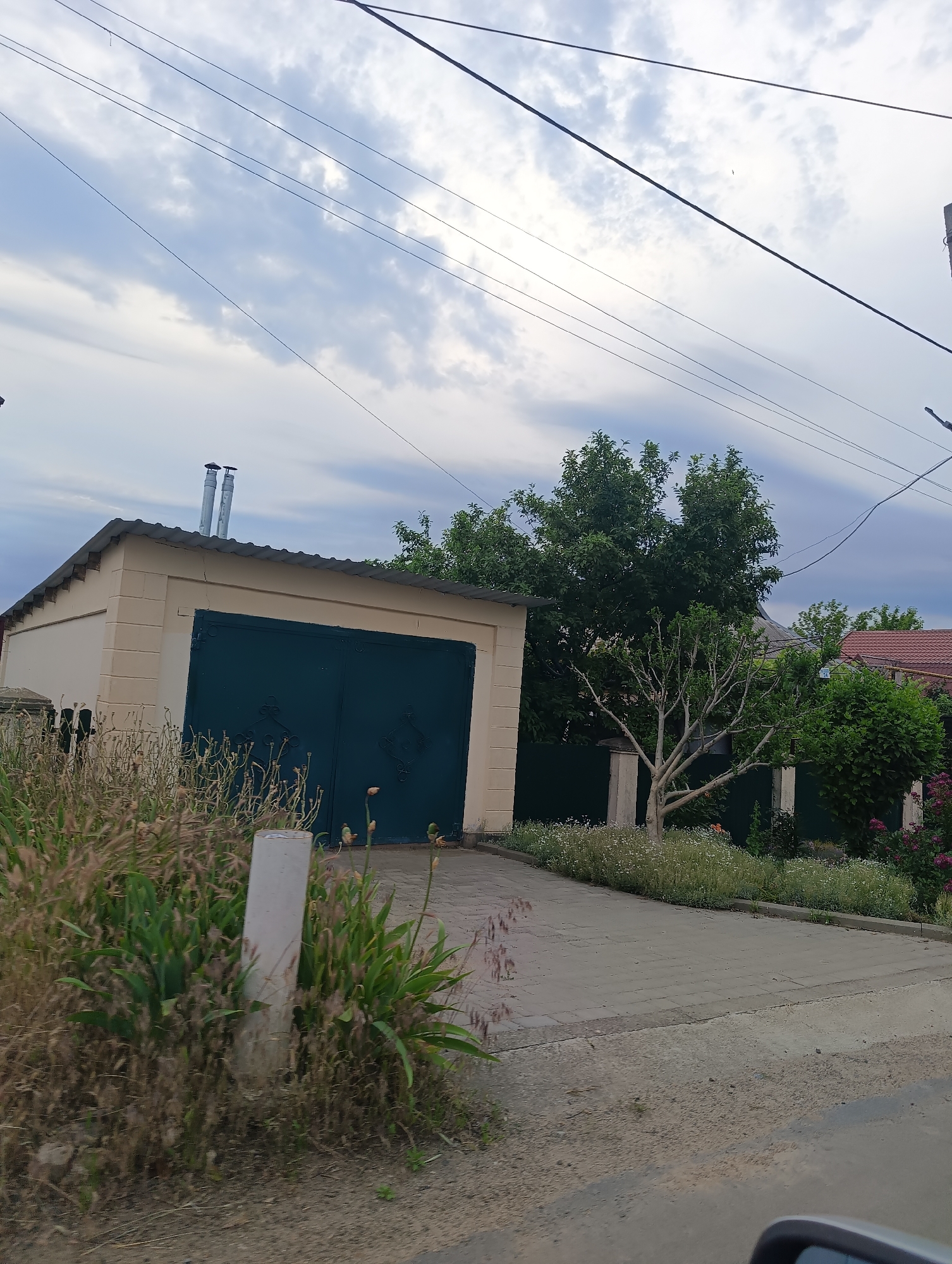 The image depicts an outdoor scene in Mayaky, Ukraine, under an overcast sky. The foreground on the left features dense, dry vegetation and green plants, with a light-colored rectangular concrete post centrally embedded within it. A dark, unpaved road surface occupies the lower right, meeting a paved driveway composed of grey rectangular blocks. The extreme lower right corner shows a partial view of a vehicle's side mirror.
Dominating the left-middle of the frame is a single-story structure, likely a garage, with light beige walls and a dark, corrugated roof. Two metallic, cylindrical pipes extend vertically from the left side of the roof. The garage entrance consists of a large, dark green double door adorned with ornate scrollwork. A fine horizontal crack is visible on the upper facade of the garage.
To the right of the garage, a landscaped area with small white flowering plants borders the paved driveway. Further right, a tree with light-colored, bare branches stands prominently, flanked by dense green foliage. A dark green fence separates the garage property from an adjacent area with more trees and a house featuring a reddish-brown tiled roof visible in the background.
The upper portion of the image is dominated by an expansive, cloudy sky with variations of light grey and white. Multiple dark power lines stretch across the sky, mostly horizontally and diagonally. A small, dark, unidentifiable object, possibly a bird, is faintly visible high in the sky, slightly to the left of the center. No individuals or specific activities are observed.