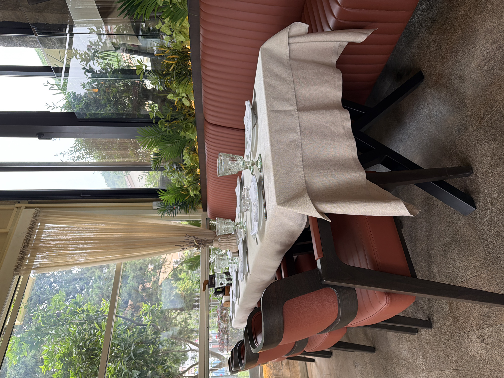 The image shows a table setting in a restaurant, likely in Budva, Montenegro. The table is covered with a light-colored tablecloth and set with glassware, plates, and silverware. It is placed along a rust-colored banquette and chairs with rust-colored cushions and dark wood frames. Large windows offer a view of lush greenery and a cityscape beyond. The overall ambiance suggests an upscale dining establishment with a focus on natural light and a pleasant view.