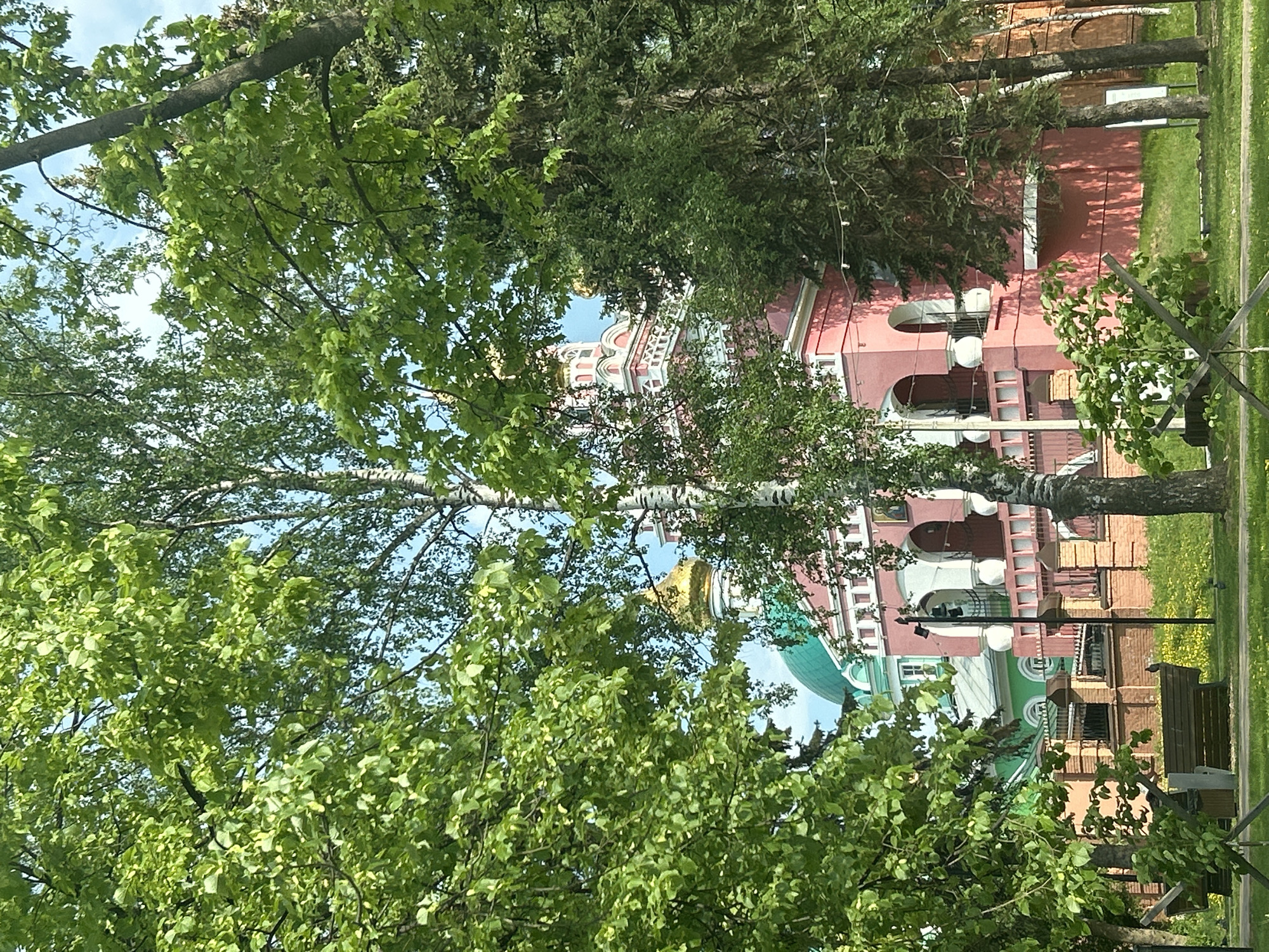 The image shows a partial view of St. Michael's Cathedral in Izhevsk, Russia, framed by trees. The cathedral is a light pink color with white architectural details, and it features green and gold domes. The scene includes a glimpse of a nearby park with a bench and path.