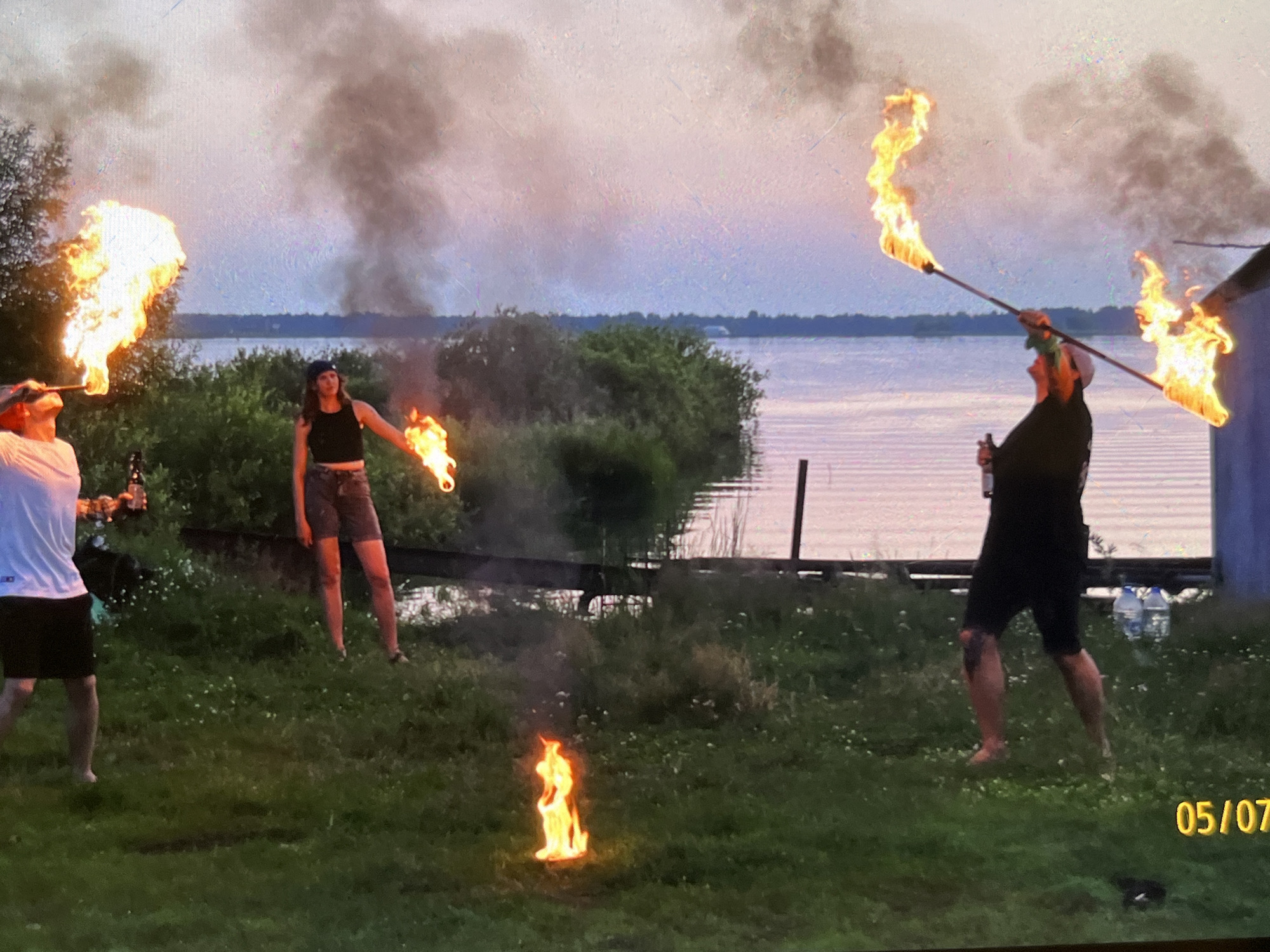 Three individuals are performing fire stunts near a body of water at dusk. On the left, a person in a white t-shirt and black shorts is blowing fire from their mouth, holding a bottle in their left hand. In the center, a person wearing a black tank top and patterned shorts stands with a lit torch in their right hand. On the right, a person in a black t-shirt and black shorts holds a long pole with two flames at either end, one directed upwards and the other downwards. In the foreground, a small fire burns on the grass. The background features a lake with a distant shoreline and a building structure on the right. The sky is a mix of gray and pink hues. The date stamp "05/07" is visible in the bottom right corner of the image. This scene does not appear to be in Tyumen, Russia.