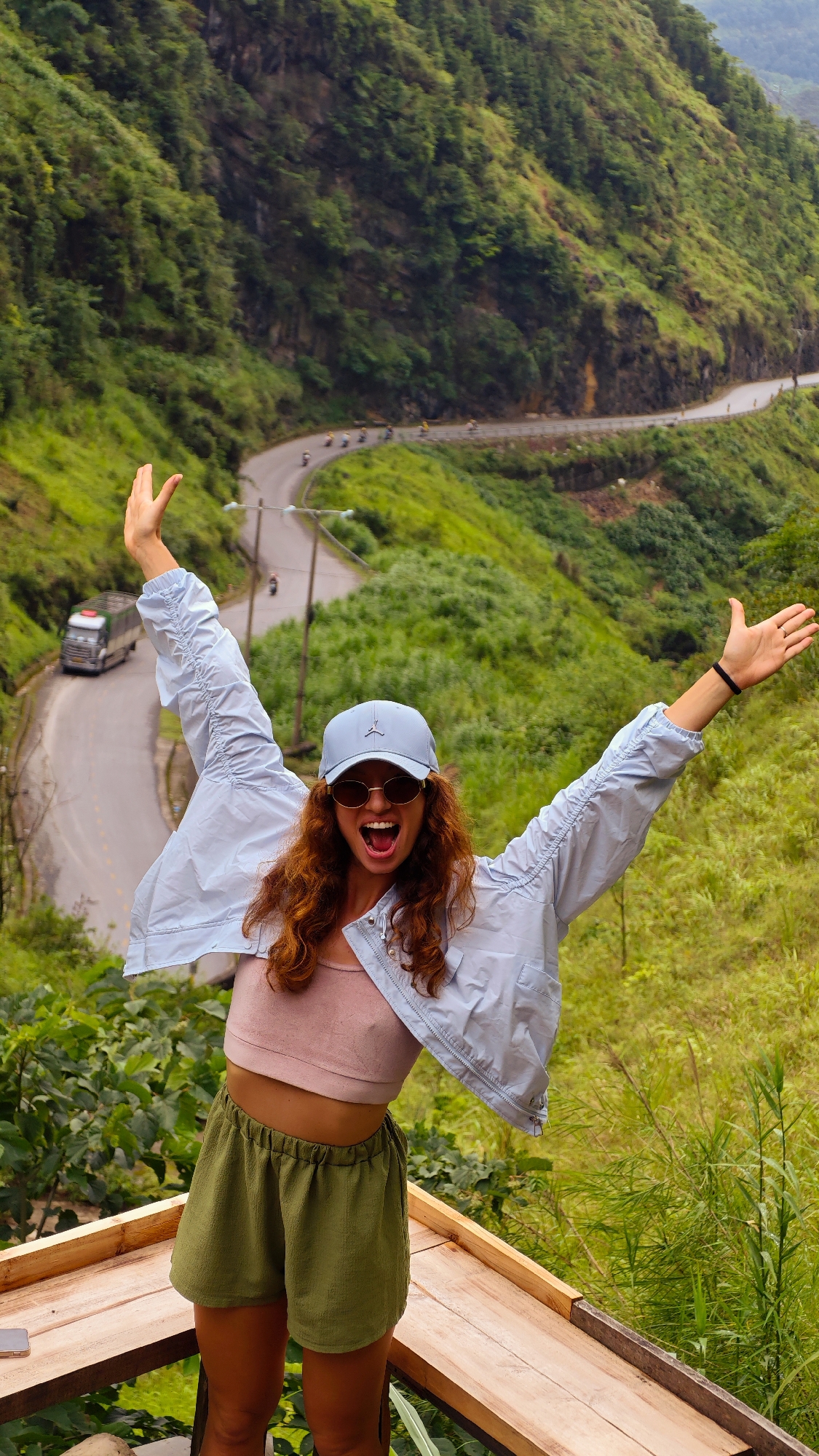 The image shows a young woman standing on a wooden platform with her arms raised in a celebratory gesture. She is wearing sunglasses, a light blue baseball cap, a light pink tank top, a light blue jacket, and green shorts. The background features a winding road with vehicles on it, lush green hills, and dense vegetation. The location is Yên Minh Commune, Vietnam, judging by the landscape.