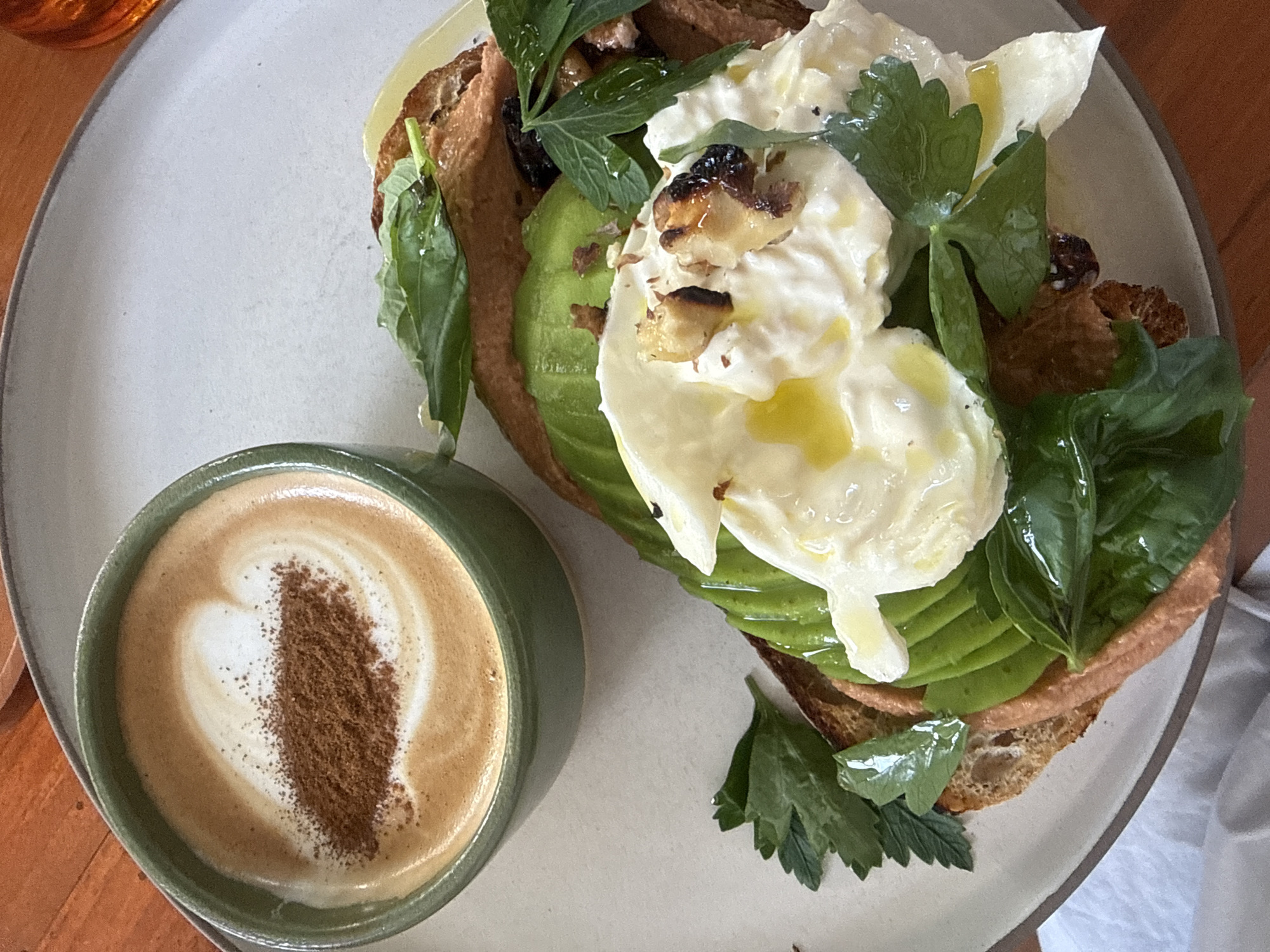 An overhead shot captures a visually appealing breakfast or brunch spread featuring avocado toast and a coffee. The main subject is a light-colored, possibly ceramic, plate holding a substantial open-faced sandwich. This sandwich consists of thick-cut toasted bread, layered with neatly sliced green avocado. On top of the avocado is a generous portion of a creamy white ingredient, resembling burrata, ricotta, or a poached egg, garnished with various fresh green herbs like parsley and basil. Scattered over the creamy topping are small, dark, caramelized pieces, possibly roasted nuts or garlic, and a glistening drizzle of olive oil.
Adjacent to the plate, positioned in the lower left, is a green ceramic mug filled with a coffee drink, likely a latte or cappuccino. The coffee exhibits intricate latte art, forming a heart shape, which is further enhanced by a dusting of cinnamon or cocoa powder down its center.
The setting appears to be indoors, with a warm, natural light illuminating the scene. A wooden surface, possibly a table, is visible in the background, providing a rustic contrast to the food. The image is bright and clear, highlighting the textures and vibrant colors of the ingredients. No text is visible in the image.