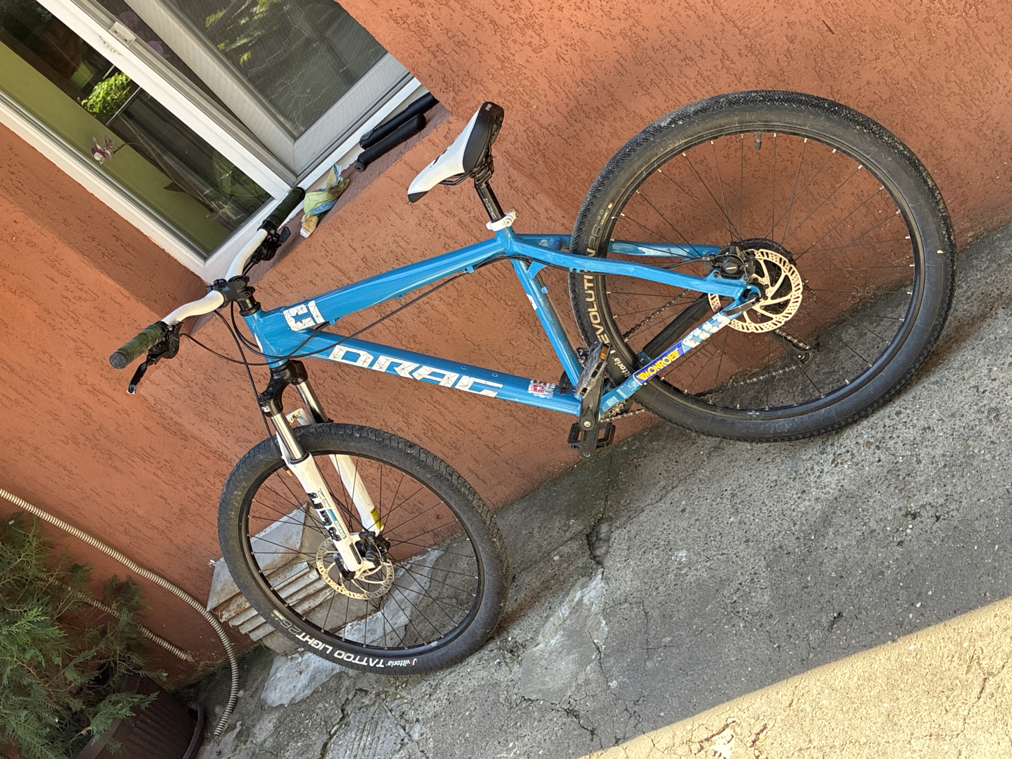 A blue dirt jump or mountain bike is prominently featured, leaning diagonally against an orange-red textured wall outdoors. The bike is the primary subject, illuminated by strong sunlight that casts distinct shadows on the concrete ground.
The bike's frame is a bright blue, displaying the brand name "DRAG" in white letters on the downtube. "EVOLUTION" is partially visible on the top tube and also on the side of the rear tire. It features white handlebars with green grips, a white suspension fork (with "RST" visible), and a white seat on a black seat post. Both the front and rear wheels are equipped with disc brakes, the rear rotor having a distinctive gold hue. The black tires show visible white lettering; the front tire reads "Vittoria TATTOO LIGHT 26", while the rear tire also shows "EVOLUTION". A "MONROE" sticker is partially visible on the bike's chainstay.
The background includes a white-framed window with a screen on the orange wall, reflecting green foliage. To the bottom left, green bushes or plants are visible near the wall, and a corrugated pipe runs along the base of the wall. The ground is made of concrete, showing cracks and a varied texture, with a stark shadow line indicating bright sunshine. No human activity is depicted; the bike is stationary. The setting appears to be a residential or urban outdoor area on a clear, sunny day, likely in Varna, Bulgaria.