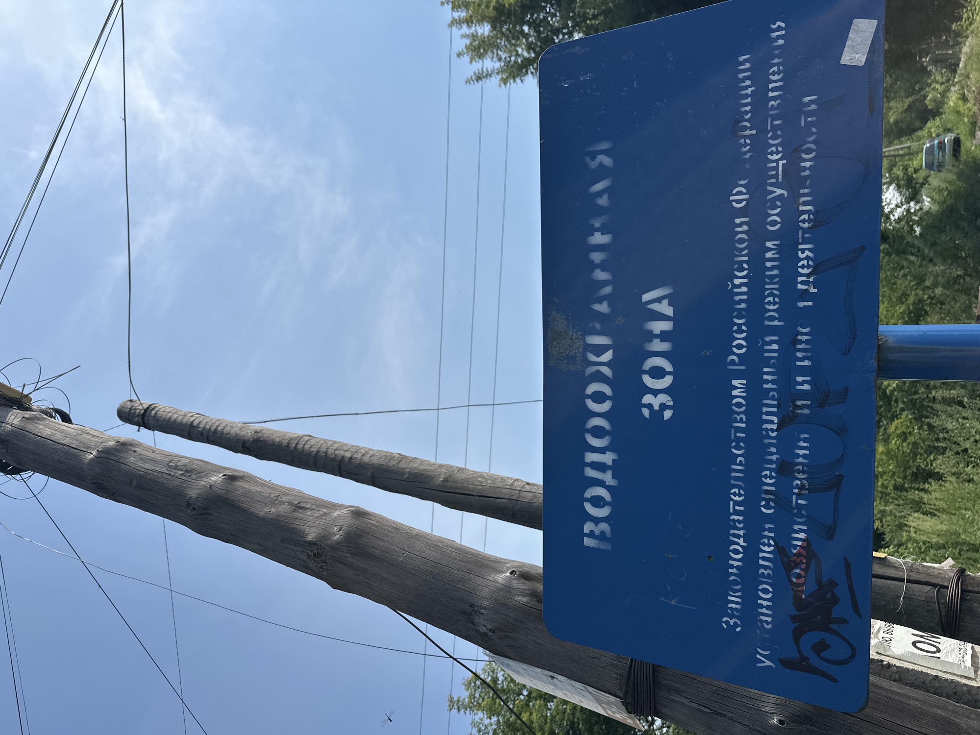 This is a medium shot, eye-level, outdoor photograph taken on a sunny day with a bright blue sky and scattered white clouds. The main subject is a blue sign attached to a weathered wooden pole. The sign has white stenciled text that reads "ВОООХРАННАЯ ЗОНА" (Water Protection Zone) and "Законодательством Российской Федерации установлен специальный режим осуществления хозяйственной и иной деятельности" (By the legislation of the Russian Federation, a special regime for conducting economic and other activities is established). The text is partially obscured by graffiti and some dirt.
The wooden pole is rough and grey, with visible grain and a crack running down its length. More wooden poles and overhead wires are visible in the background, suggesting an outdoor utility area. Green trees and foliage form the background landscape. In the lower left corner, another white sign is partially visible with some text. A small, dark dragonfly is in flight to the left of the main pole. In the distance, a light-colored vehicle is parked near the trees.
The image location is identified as Barnaul, Russia, based on the Cyrillic text on the sign. The overall impression is of a functional, outdoor sign in a somewhat rural or semi-rural setting within the city, possibly near a body of water due to the "Water Protection Zone" designation. There are no people visible in the photograph.