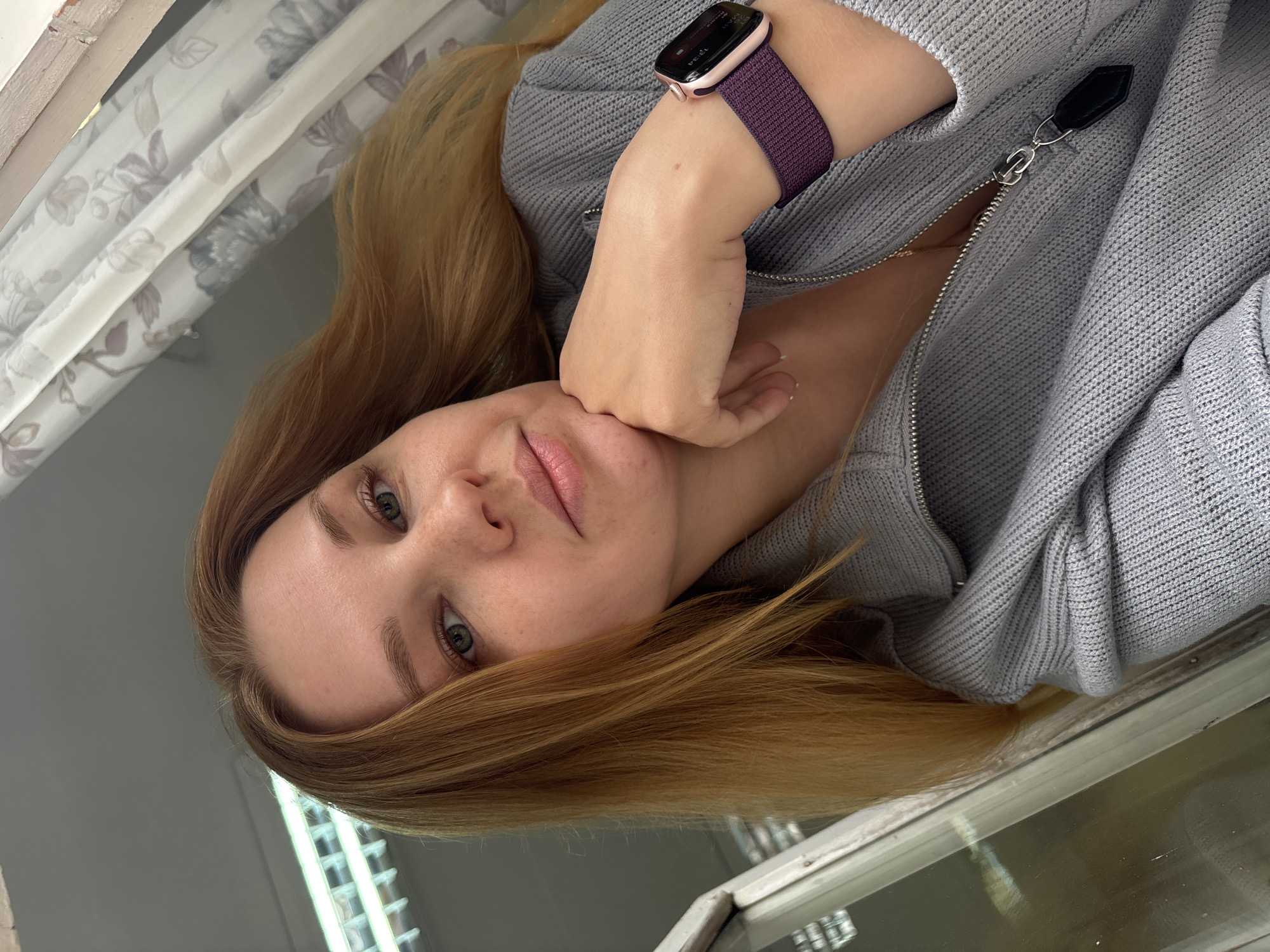 A woman with light skin, light eyes, and reddish-blonde hair is the primary subject of the image. She is wearing a gray, knitted zip-up sweater and an activity tracker with a purple band on her left wrist. Her hand rests on her face. The scene is indoors, near a window with floral patterned curtains, and a reflection is visible in the windowpane. Rusanovo, Russia cannot be verified from the provided image.
