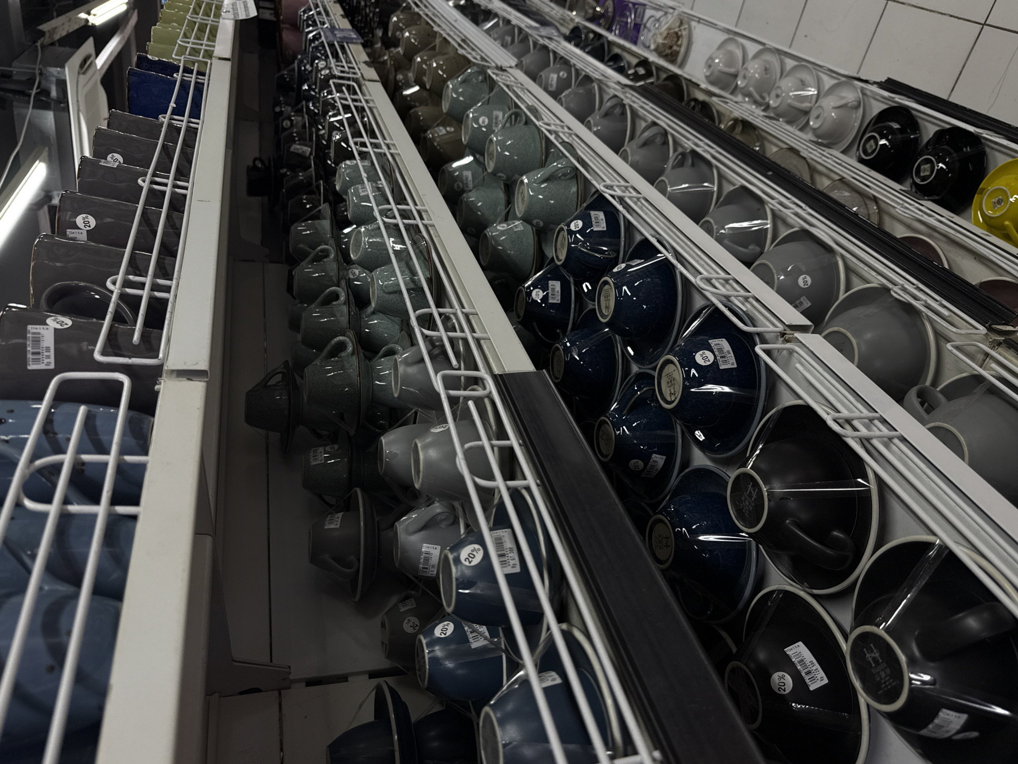 The image shows multiple shelves containing a variety of ceramic cups in a store setting. The cups are arranged in rows and columns, with different colors ranging from gray to blue and black. White wire shelving supports the cups. The scene is brightly lit, likely from the store's overhead lighting. The store might be located in Ubud District, Indonesia. Labels on the cups display price and discount information, indicating a retail environment.