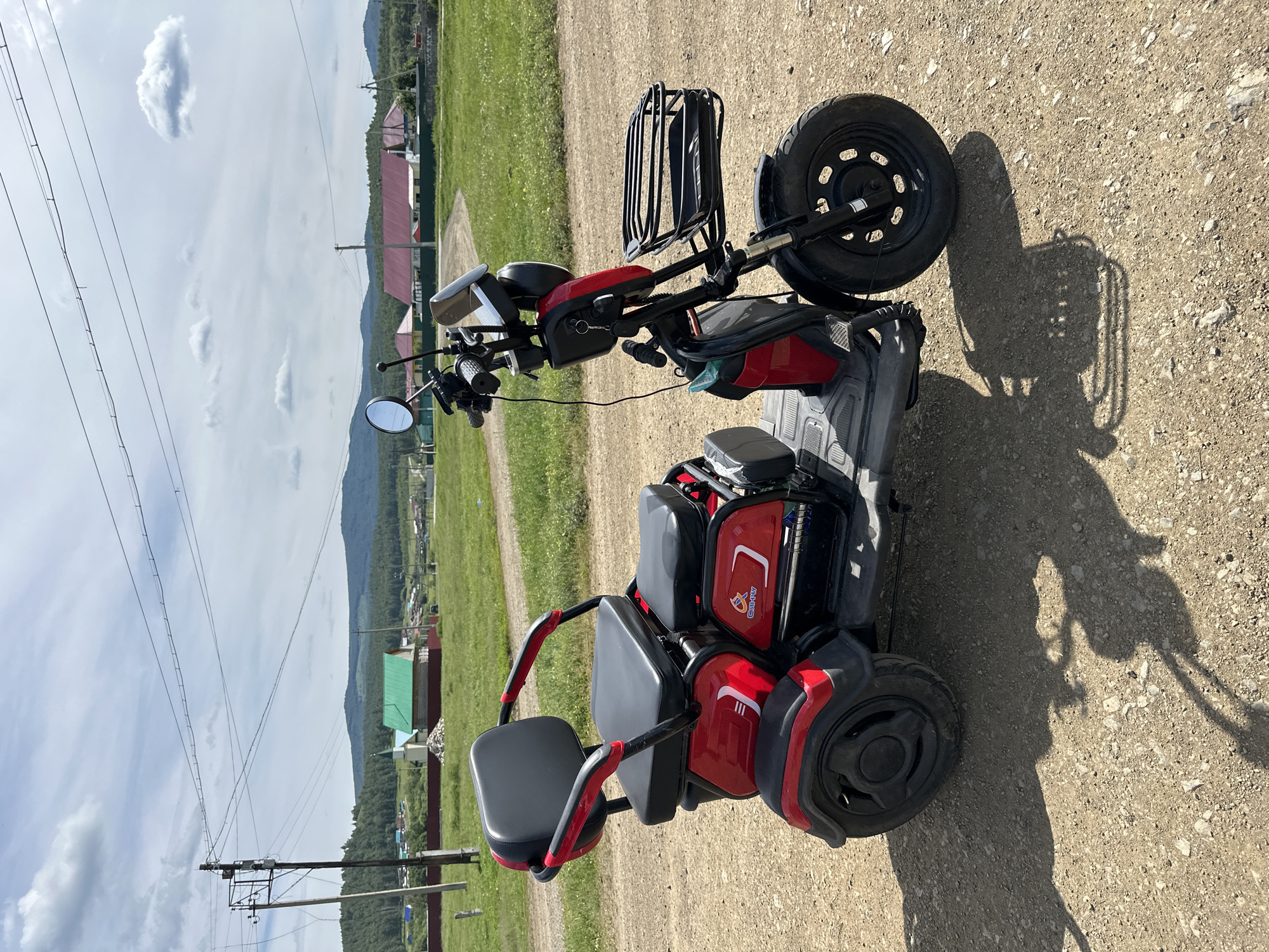 An eye-level, full outdoor shot captures a red and black three-wheeled electric scooter parked on a dirt road. The scooter is oriented with its front facing right, and its right side is fully visible. It features a black seat with armrests, a front basket, and a black handlebar. Behind the scooter, a rural landscape unfolds under a cloudy sky. Green hills covered in trees form the horizon, with a village of houses featuring red and green roofs nestled in the middle distance. A power line pole with wires stretches across the top of the frame. The ground is composed of dirt and small stones, casting distinct shadows from the scooter.