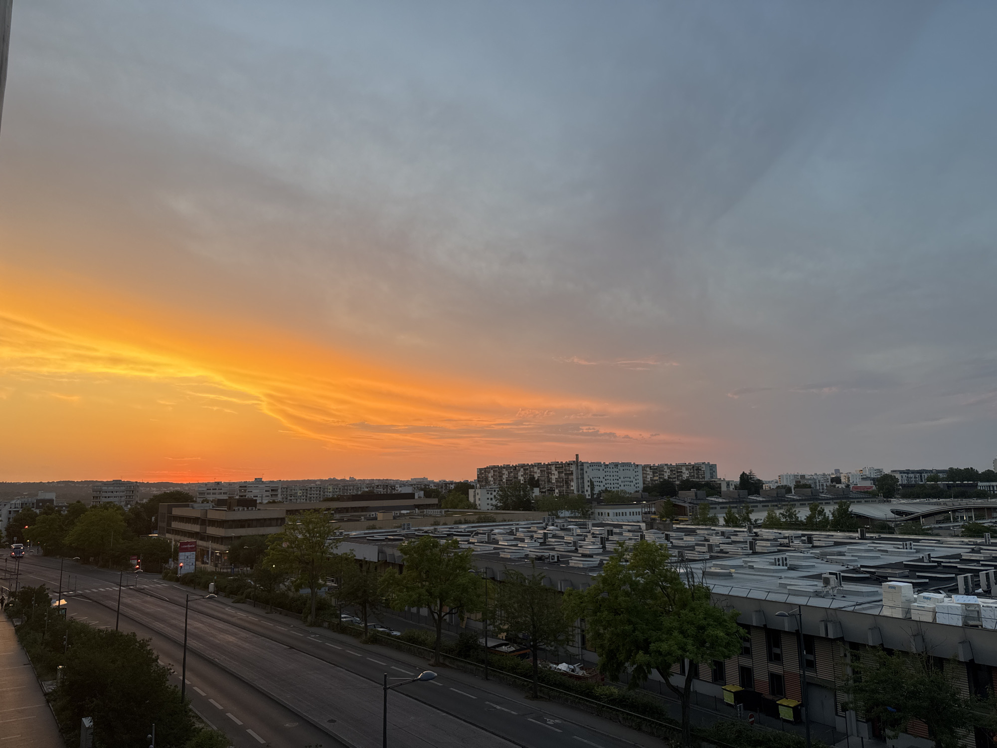 An aerial, wide-angle, full shot captures a cityscape at dusk with a vibrant, orange-hued sunset dominating the left half of the frame and a muted, blue-gray sky on the right. The sky transitions from intense orange near the horizon to a lighter orange and then to a pale blue-gray with subtle cloud formations. Below the sky, an urban landscape unfolds, featuring rows of apartment buildings, some with balconies, and several low-rise commercial or industrial buildings with flat, white-topped roofs that appear to be equipped with HVAC units. A tree-lined street with multiple lanes of traffic runs diagonally across the foreground, with a few cars visible. The street is illuminated by streetlights, and sidewalks are present on both sides. The overall scene suggests a residential and commercial area in a city experiencing sunset.