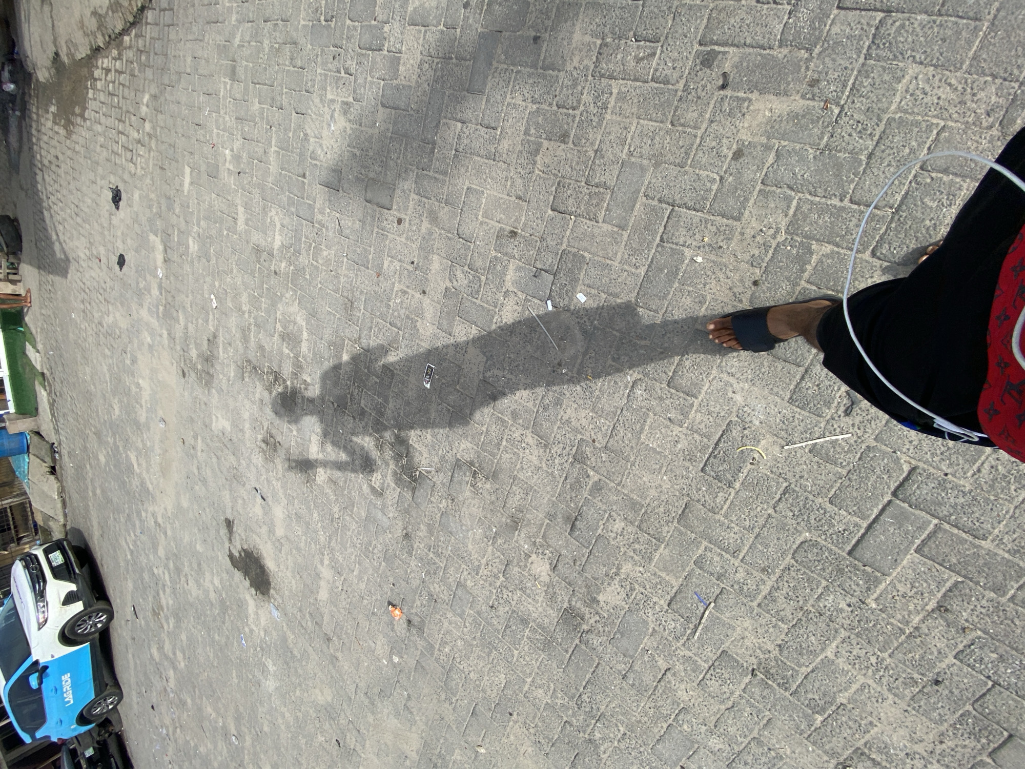 The image captures an outdoor scene in Ado, Nigeria, on a paved area constructed of interlocking concrete blocks. The photograph is taken from a slightly elevated perspective, looking down.
Visible is the shadow of a person, likely the photographer, holding a phone or similar device. To the right, a person's legs and feet are visible, clad in black trousers and one foot in a dark sandal. Part of a red patterned bag is also in the frame. A white wire, possibly from headphones, is present.
A LagRide car is parked to the left of the frame.
Debris, including small pieces of litter, is scattered across the pavement.