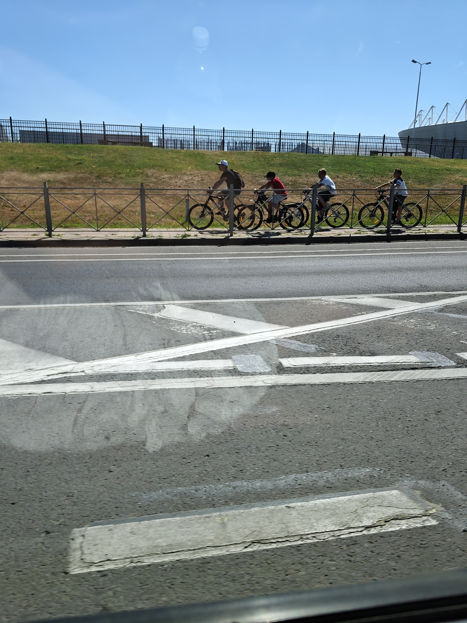 This image captures a sunny day in Rostov-on-Don, Russia, with a clear blue sky overhead. In the middle ground, a group of four individuals are riding bicycles along a paved path. The first cyclist, wearing a cap, is on the left. To their right, three younger individuals are riding their bicycles in single file. They are separated from the road by a metal fence with a diamond pattern. Beyond the path and fence is a grassy embankment, also secured by a black metal fence. In the background, a modern stadium with a white, angular roof is visible, along with a tall lamppost. The foreground of the image shows a section of asphalt road with white markings, possibly indicating a pedestrian crossing. The image appears to have been taken from a vehicle, with a slight reflection visible on the left side.