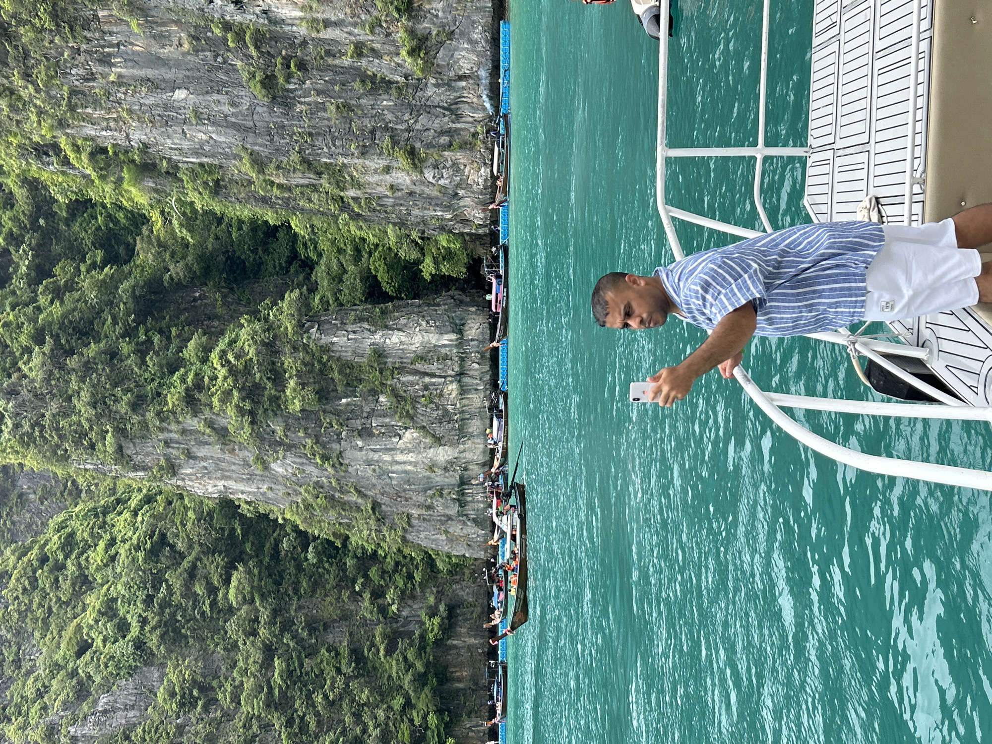 In Ao Nang, Thailand, a man stands on the deck of a boat, holding a white smartphone in his right hand, appearing to take a photo of the surrounding scenery. He is wearing a blue and white striped short-sleeved shirt and white shorts. The boat is on a body of turquoise water, with several long-tail boats visible in the distance. Behind the boats, a dramatic cliff face covered in lush green vegetation rises steeply from the water. The sky is overcast.