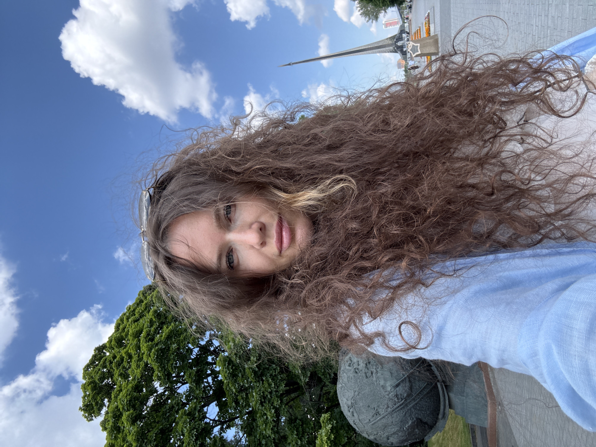 A woman with long, curly brown hair takes a selfie outdoors in Moscow, Russia. She is wearing a light blue top. Behind her, a large green tree is visible on the left, and a monument, possibly the Monument to the Conquerors of Space, stands on the right. The sky is blue with scattered white clouds. A large, globe-like sculpture is situated to the left of the woman. The ground is paved with light-colored bricks.