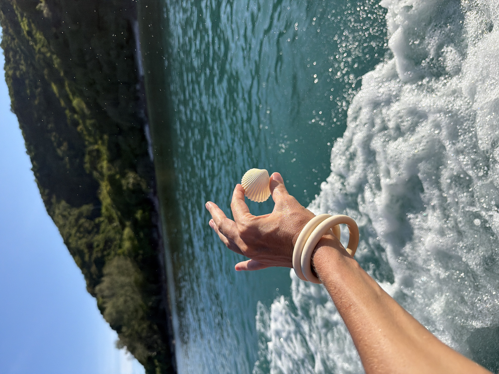 The image shows a hand holding a white seashell, with two light-colored bracelets on the wrist. The hand and arm are positioned in the foreground, in front of a turbulent, turquoise body of water, likely the sea, with white foam and spray. In the background, a steep, tree-covered incline rises from the water, with a clear blue sky above. The lighting suggests it is daytime, likely sunny, given the bright reflections on the water and the clarity of the sky. The overall scene evokes a sense of vacation and seaside exploration, possibly from a boat, given the movement of the water. The location context points to Baan Thong Krut, Thailand, a known area for beaches and coastal activities. No text is visible in the image.