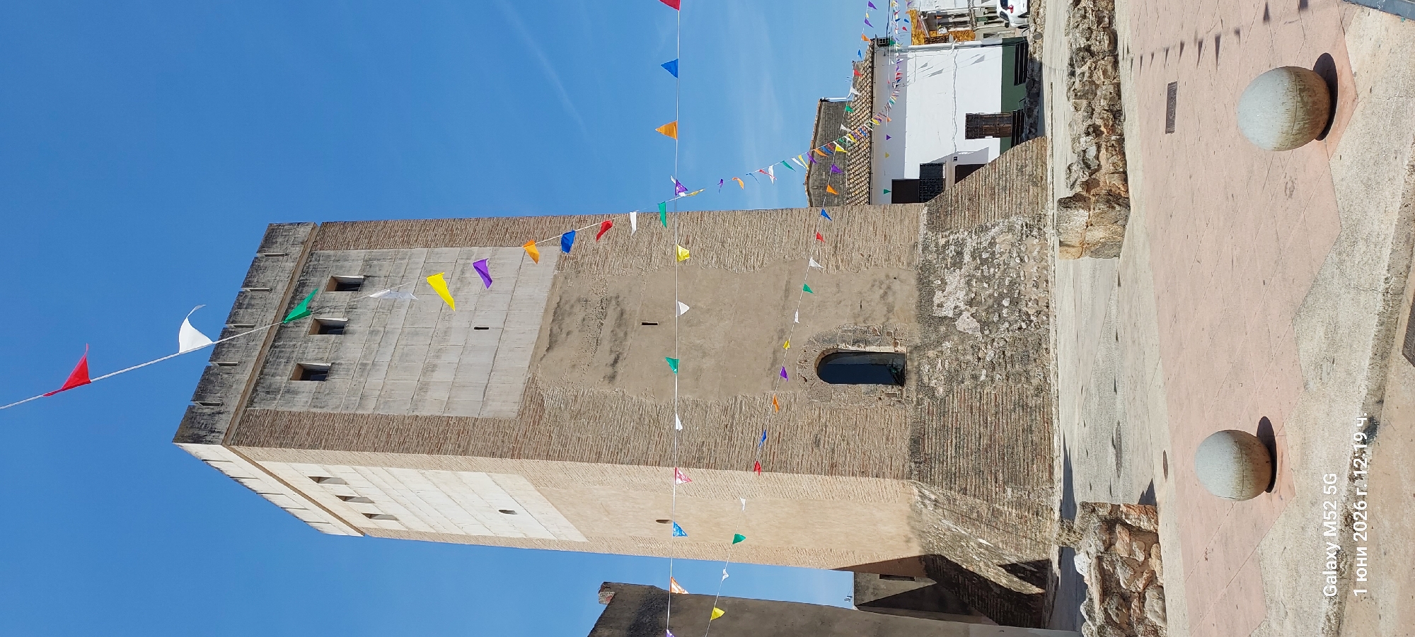 The image depicts a brick tower decorated with colorful triangular flags. The tower features a mix of brick and cement construction, with several windows visible. The sky is clear and blue. The surroundings include other buildings with white walls and tiled roofs. In the foreground, there is a stone path with spherical stone bollards. The image appears to be taken at ground level, looking upwards at the tower. The image's timestamp indicates it was taken on June 1, 2026. The city of Canals, Spain, is identified as the location.