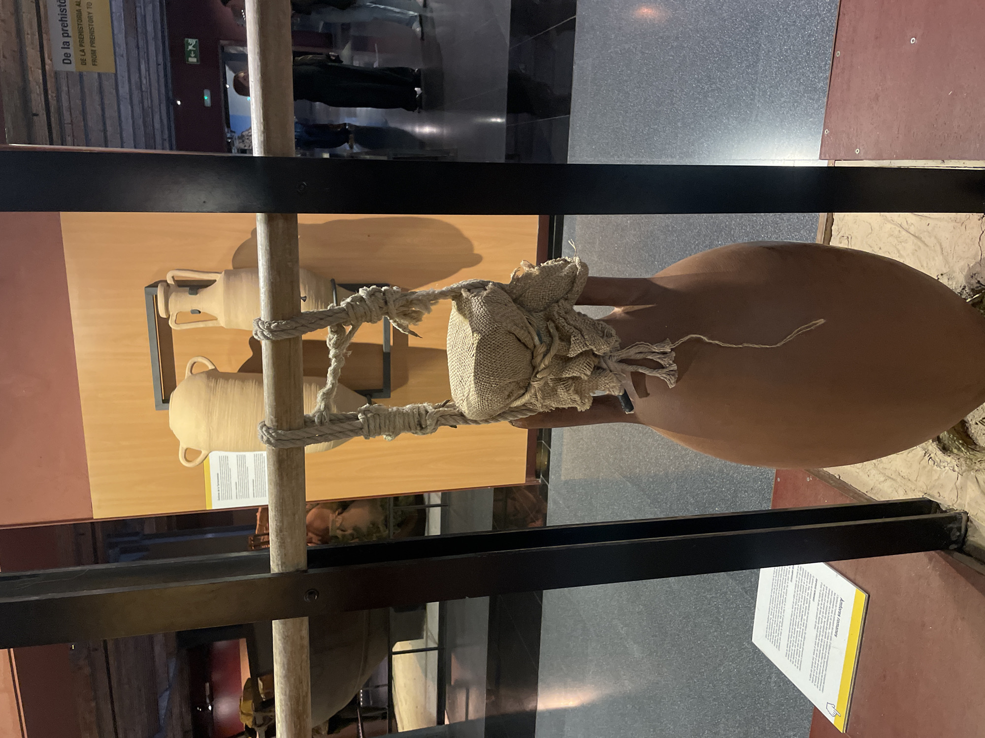 The image depicts an indoor museum exhibit, likely in Barcelona, Spain, showcasing ancient ceramic vessels. The primary focus is a large, reddish-brown amphora, positioned on the right side of the frame. This amphora is supported by thick, natural fiber ropes that are intricately knotted around its neck, and it appears to be further held in place by a wooden pole running vertically in front of it. A small, light-colored burlap or coarse fabric sack is tied around the amphora's neck, possibly for protection or decorative purposes.
In the background, to the left, another lighter-colored amphora with two handles rests on a wooden shelf, casting a shadow. The exhibit features black metal beams as part of its structural framework, with a wooden pole extending through the center. The floor beneath the main amphora transitions from a dark grey/black to a reddish-brown section, and a lighter, sandy-textured area is visible under the amphora's base.
Blurred reflections on glass surfaces reveal indistinct figures of museum visitors and other parts of the exhibition hall, indicating artificial indoor lighting. Visible text on a yellow sign in the bottom right corner clearly identifies the artifacts as "Anforas romanas" (Roman Amphorae), with additional, smaller explanatory text below. Another partial yellow sign in the upper left background contains the words "De la prehistoria" and "FROM PREHISTORY TO", suggesting the broader theme of the exhibit. The overall scene captures a moment of viewing historical artifacts in a well-organized museum setting.