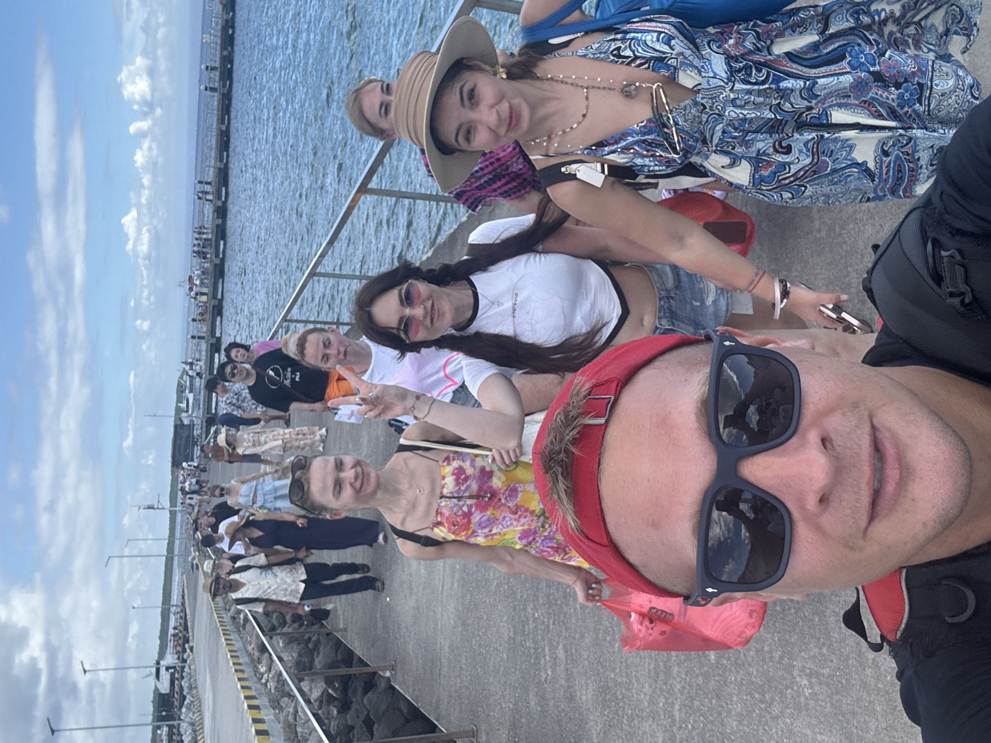 A selfie taken on a pier in Kampung Toyapakeh, Indonesia, captures a group of people. The pier stretches into the water with people walking along it, and it is lined with a metal railing. The foreground features the photographer, wearing sunglasses and a red head covering, taking the photo with a group of people standing behind them. The background shows a body of water, a cloudy sky, and the pier extending into the distance.