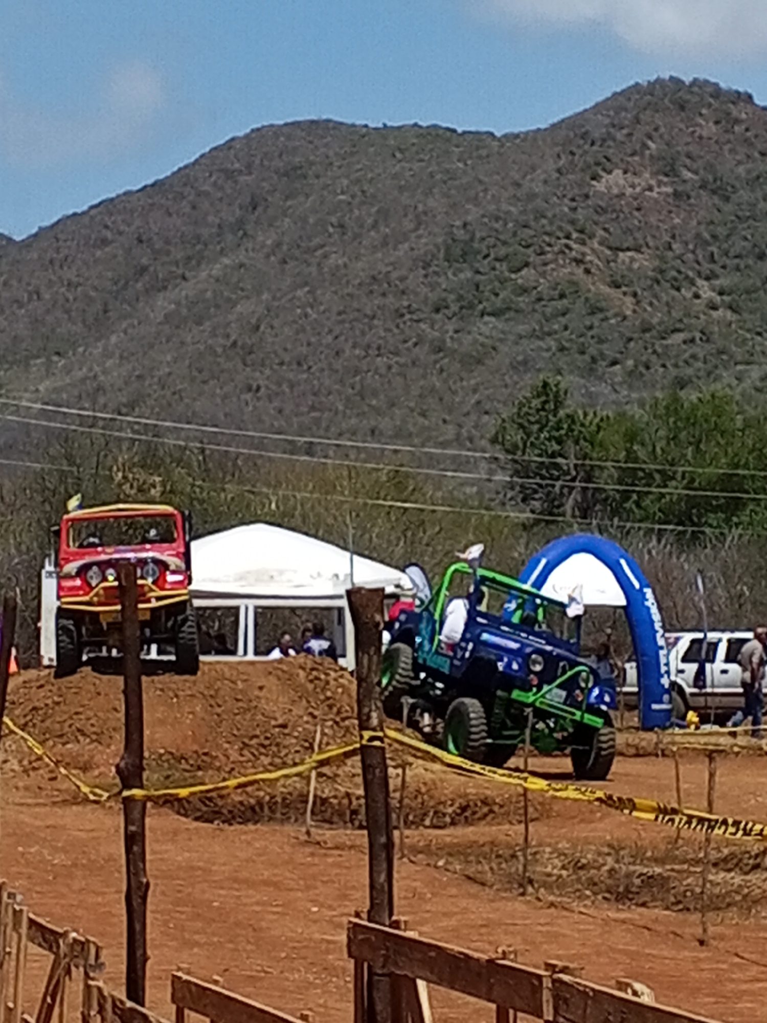 The image captures an off-road vehicle competition. A red jeep is positioned atop a dirt mound, while a blue and green jeep navigates the course. A white tent and an inflatable arch are visible in the background. The terrain is dirt, surrounded by wooden posts and yellow caution tape. A mountainous landscape forms the backdrop under a clear blue sky. The location is the city of Unknown, Unknown. A white SUV and a man can be seen near the inflatable arch. People are present under the tent.
