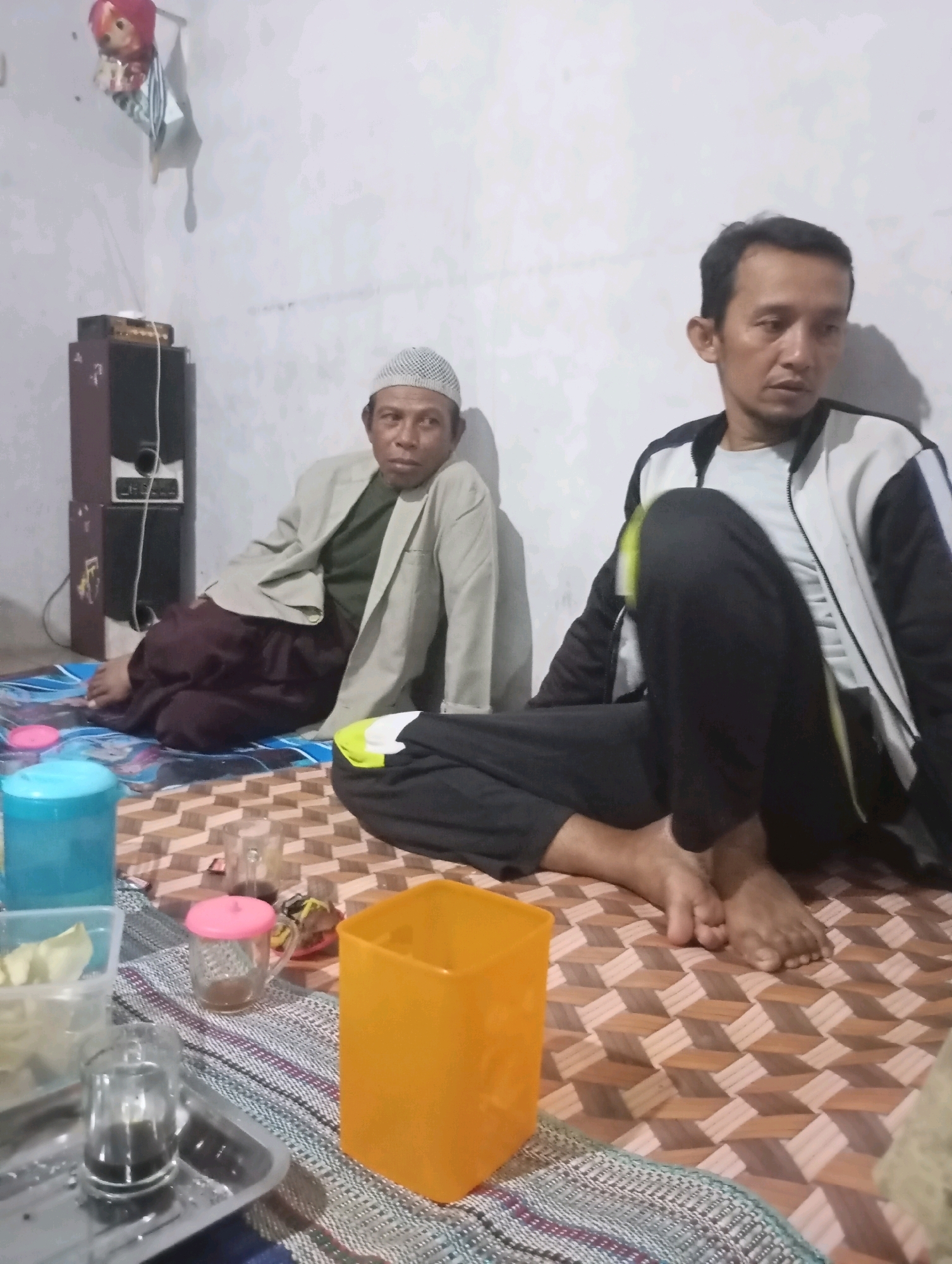 In the city of Wonogondo, Indonesia, two men are seated on a patterned mat. The man on the left wears a light brown jacket over a green shirt and maroon pants, with a white cap on his head. He is leaning against a speaker system. The man on the right wears a black and white jacket over a grey shirt and black pants. He is looking to his left. In front of them are several objects, including a blue plastic container, a clear plastic container with what appears to be food, a small glass with a pink lid and brown liquid, a metal tray with a glass of brown liquid, and an orange plastic container. The environment appears to be indoors, with a light-colored wall in the background.