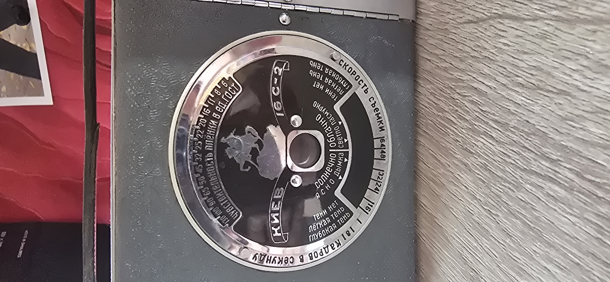 The image is a close-up, high-angle shot of a vintage camera's light meter dial. The dial is circular with a metallic silver rim. The central part of the dial is black and features a raised silver silhouette of a horseman. Text and numbers in Russian are inscribed around the dial. At the top, in a curved arc, is the text "ЧУВСТВИТЕЛЬНОСТЬ ПЛЕНКИ В ЕД. ГОСТ", which translates to "FILM SENSITIVITY IN GOST UNITS," with numbers ranging from 13 to 130. Below this, in a larger font, is "КИЕВ" (KIEV) and "16 С-2". Around the lower part of the dial, other inscriptions include "СКОРОСТЬ СЪЕМКИ" (SHOOTING SPEED), with numbers like 64, 125, 250, and also what appears to be aperture settings like 1.8, 2.8, 4, 5.6, 8, 11, 16. There are also descriptions of lighting conditions such as "СОЛНЕЧНО" (SUNNY), "ОБЛАЧНО" (CLOUDY), "ЯСНО" (CLEAR), "ДЫМКА" (HAZE), "СВЕТЛО ПАСМУРНО" (LIGHTLY OVERCAST), and descriptions of shadows like "ТЕни нет" (NO SHADOWS), "легкая тень" (LIGHT SHADOW), "глубокая тень" (DEEP SHADOW). The dial is set against the dark gray, textured body of the camera. The background is out of focus and appears to be a red, textured surface, possibly fabric, with a sliver of a white-framed picture visible at the very top. The camera is resting on a surface that appears to be made of wood with a textured grain pattern. The image is well-lit, highlighting the details of the camera's dial. There is no indication of the city of Gostagayevskaya, Russia, in the image.