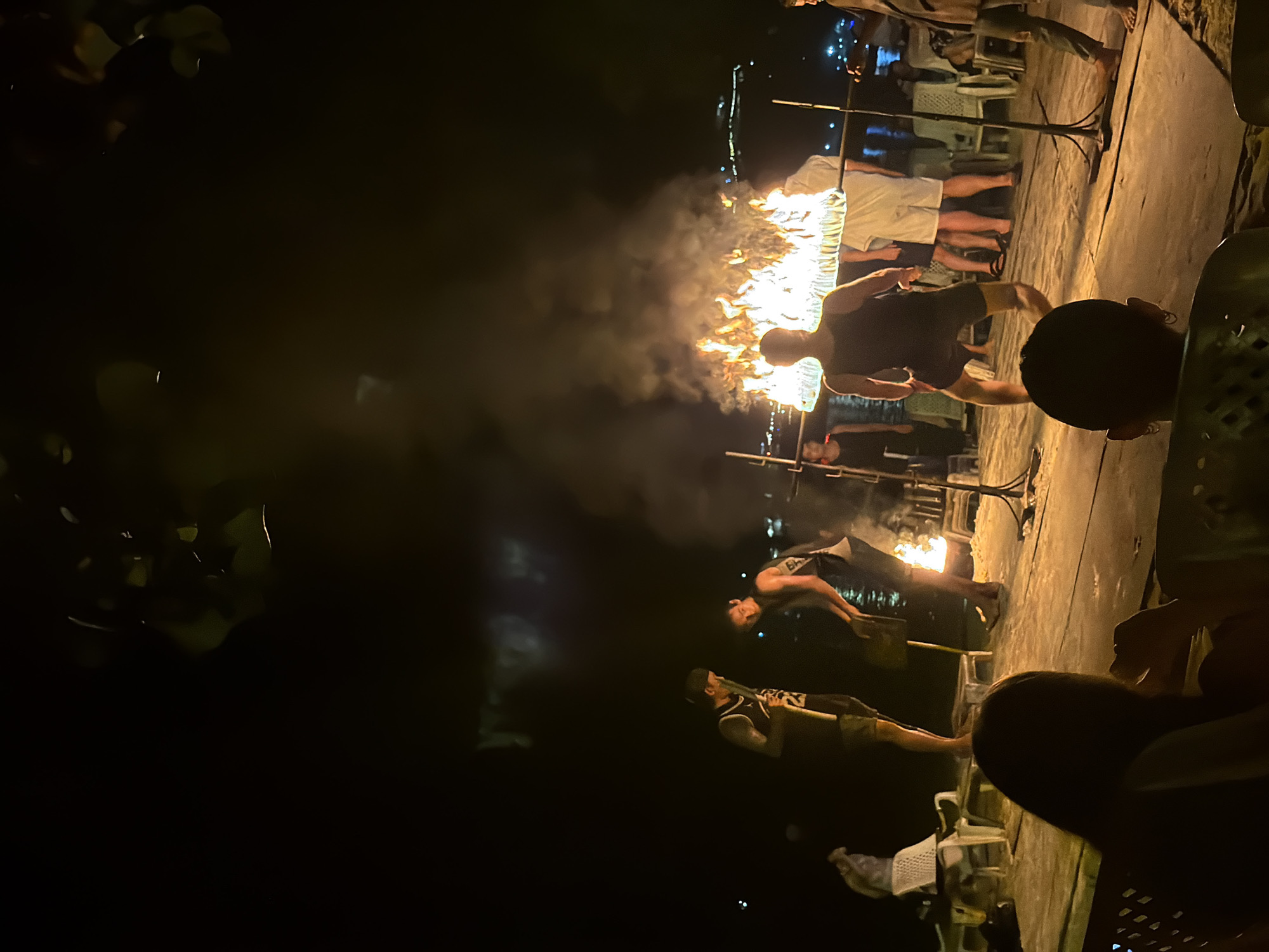 In Ao Nang, Thailand, at night, a group of people are gathered around a large, horizontal fire display that is suspended by metal poles. The fire illuminates the immediate area, creating a dramatic scene against the dark night sky. Several individuals are positioned near the fire, some appearing to be performers or involved in its operation. In the foreground, the backs of two spectators are visible, seated and watching the spectacle. In the background, the lights of a town or resort are visible across the water, suggesting a beachfront or waterfront location. The ground appears to be sand or a similar beach material. Several chairs, likely for an audience, are scattered around the area.