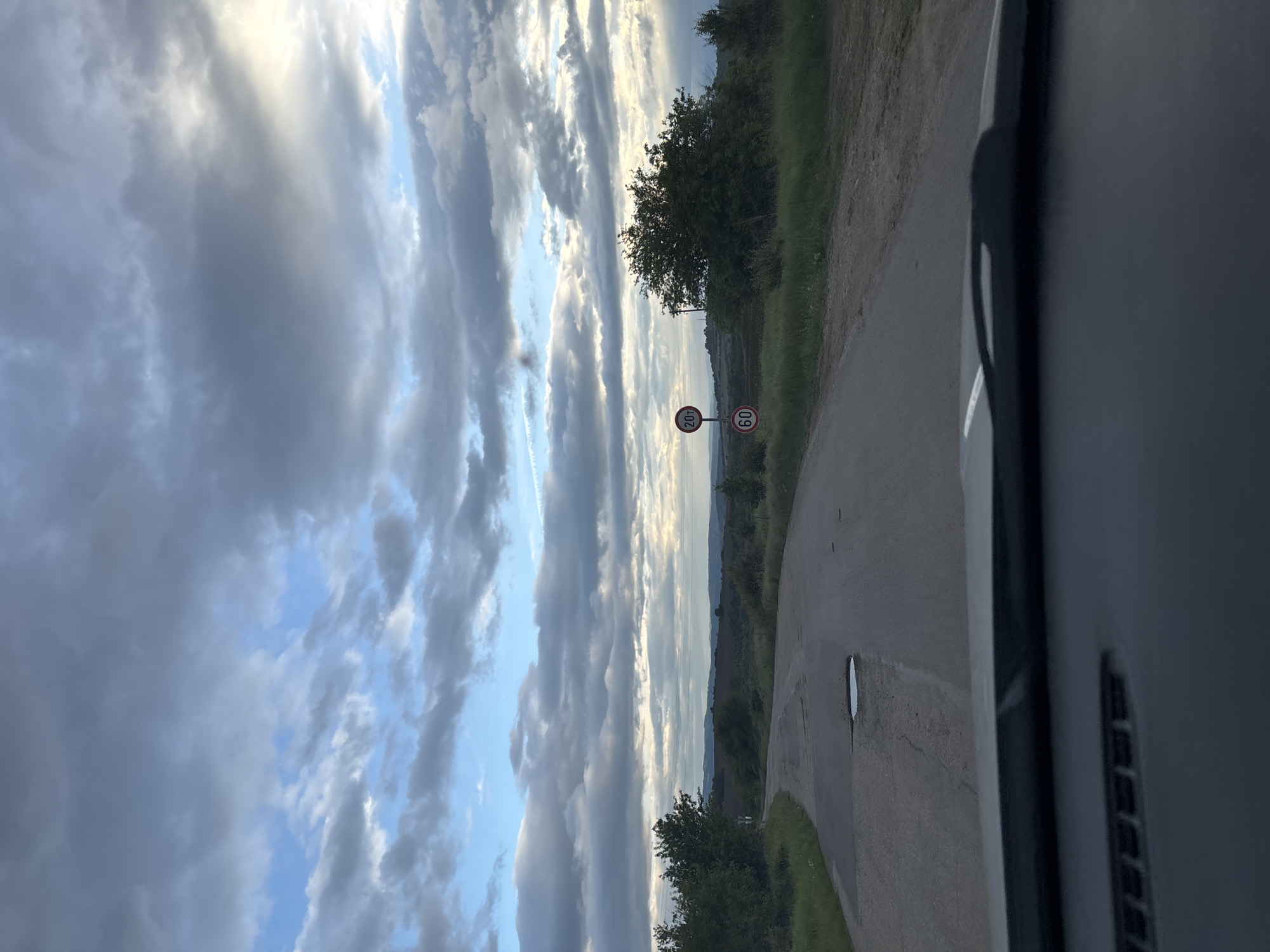 The image depicts a view from inside a vehicle, likely a car, on a rural road in the vicinity of Baylovo, Bulgaria. The road curves to the right. A speed limit sign indicating "60" and another sign stating "20t" (likely a weight restriction) are visible. The sky is filled with a mix of clouds and patches of blue. Vegetation, including trees and grass, lines the side of the road. The car's dashboard is visible in the foreground.