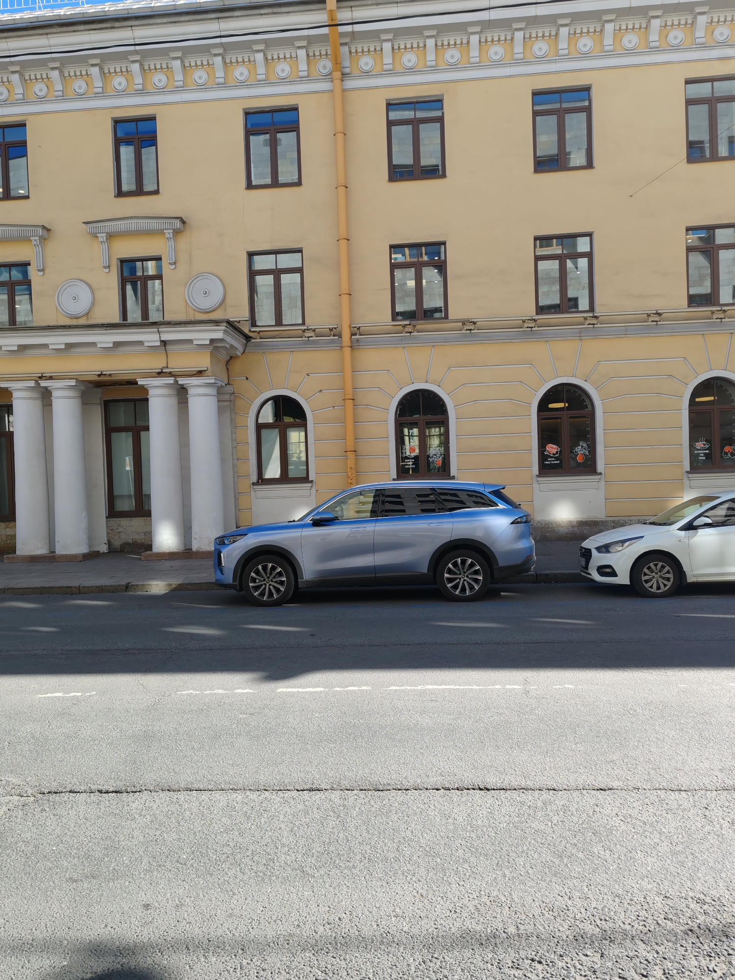 This daytime image showcases a street scene in Saint Petersburg, Russia, featuring a grand, light yellow multi-story building with classical architecture. The building's facade is adorned with white decorative cornices, moldings, and prominent white columns forming an entrance on the left. Numerous windows with dark frames are visible: arched ones on the ground floor and rectangular ones on the upper levels. A cream-colored pipe runs vertically along the right side of the building.
Two cars are parked parallel to the curb: a sleek, light blue or silver SUV with distinctive multi-spoke wheels, and to its right, a partially visible white sedan, likely a Hyundai Solaris/Accent. The asphalt road in the foreground is dry, exhibiting a horizontal crack and a faded white dashed line. Bright sunlight casts shadows, suggesting a clear day, either morning or afternoon.
No people are visible in the scene. Several circular decals are affixed to the ground-floor arched windows. These decals feature red and orange designs, some with stylized graphics of food items such as a cut of meat, fruits, or prepared dishes. Faint text is present on some decals; given the location, this text is in Russian. One decal on the far right window appears to read "ДОСТАВКА НА ДОМ" (Delivery to home), indicating a potential business on the ground floor. The overall impression is one of a quiet, sunlit urban street.