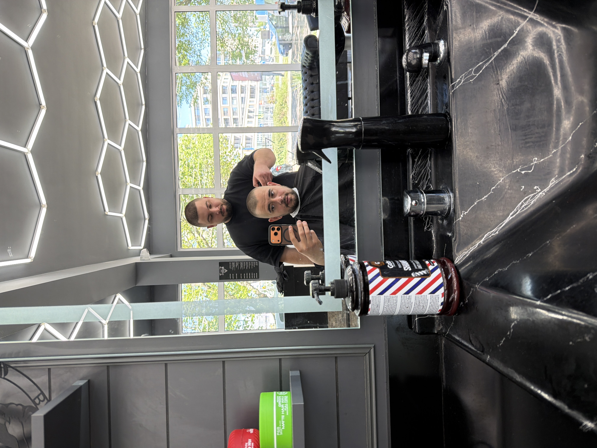 The image shows a man getting a haircut in what appears to be a barber shop. The man is wearing a black barber cape and holding a phone to take a selfie in the mirror. The barber is wearing a black t-shirt and holding a trimmer to the man's hair. On the wall behind them, there is a window with a view of buildings and trees. Below the mirror is a shelf with grooming products. The room has a modern design with a honeycomb pattern light fixture. The city is unknown.