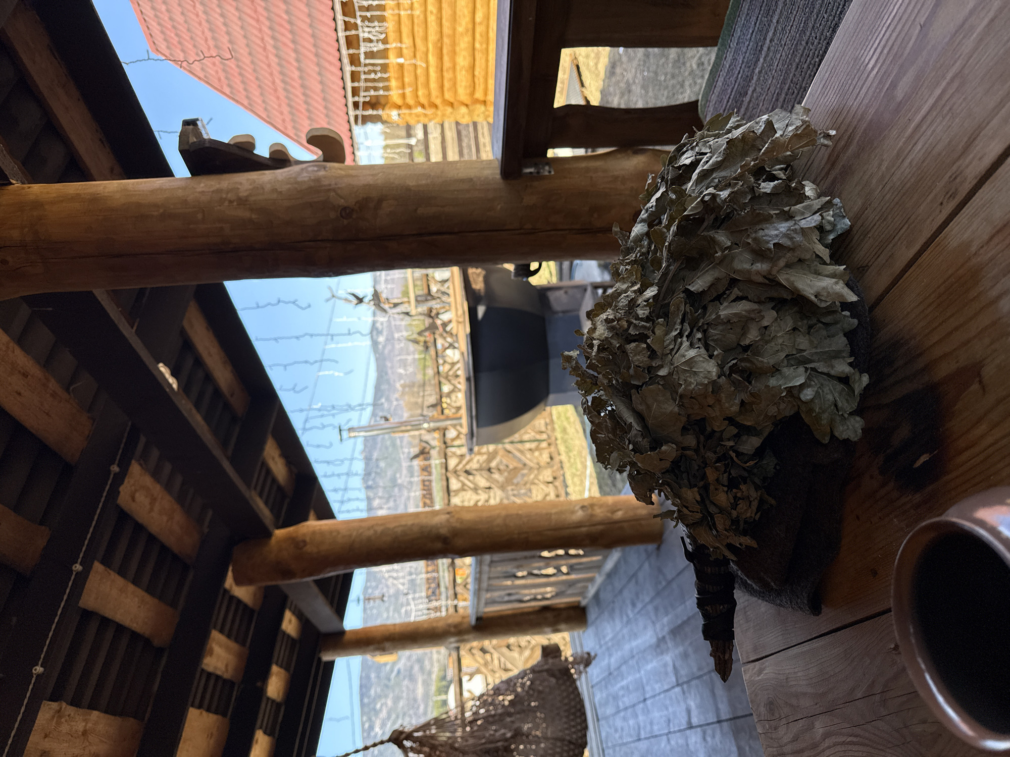 The scene is a view from inside a structure in Tyulyuk, Russia, likely a sauna or similar facility. The image is taken at an angle, looking upwards and outwards through the building's frame. The construction consists of wooden beams and planks. Through the structure, a distant landscape with hints of mountains and a sky is visible. On a wooden surface in the foreground, there is a bundle of dried birch leaves (a "venik") and a copper mug. The overall impression is rustic and suggestive of a traditional bathing or wellness experience.