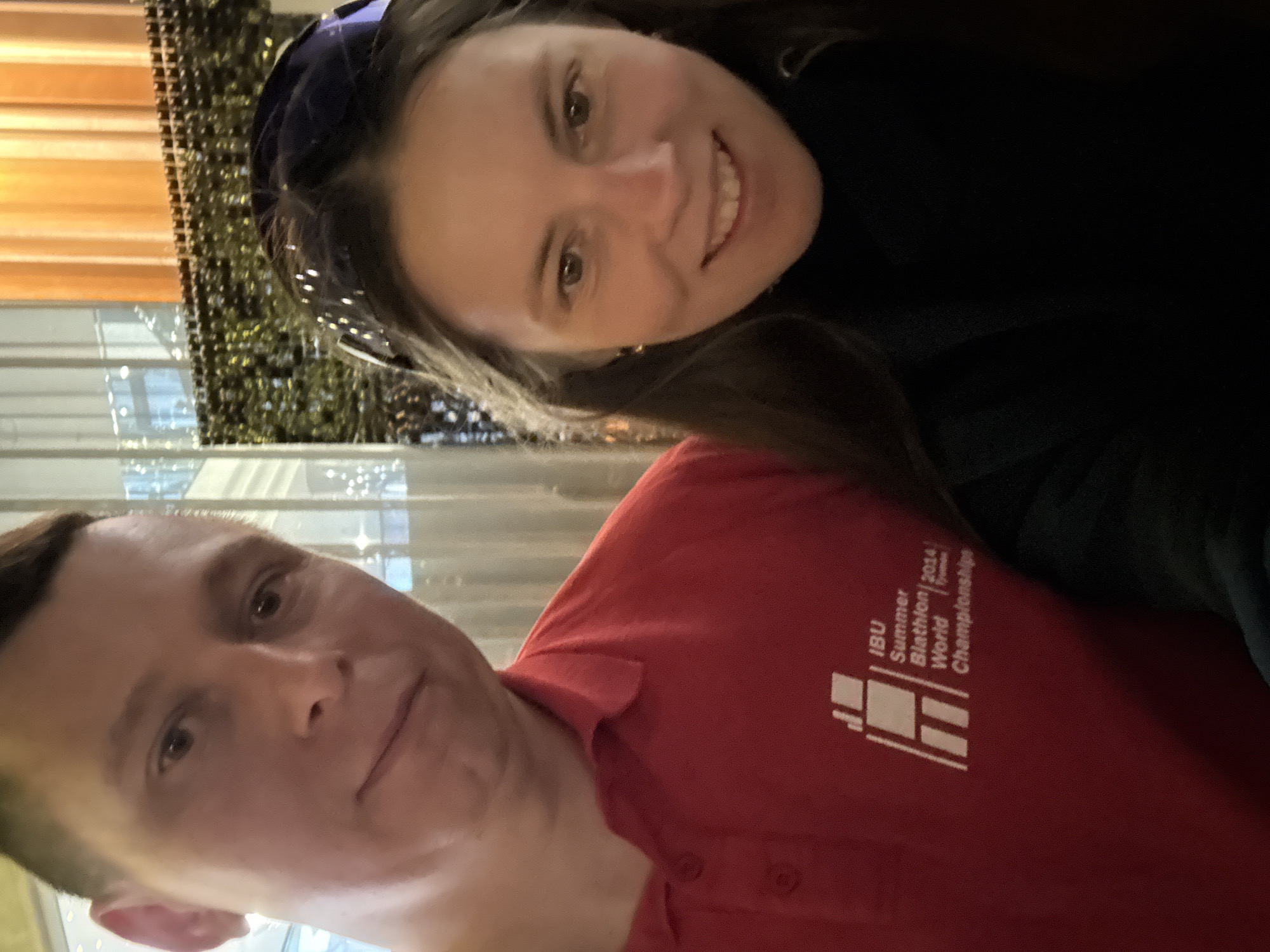 This media file captures two individuals, a man and a woman, posing closely for a picture, likely a selfie, in an indoor setting.
The woman, positioned on the right side of the frame, has long brown hair, dark eyes, and fair skin. She is smiling broadly at the camera, showing her teeth, and her expression conveys happiness and warmth. She wears a dark-colored top and a dark blue or purple headpiece.
The man, positioned to the left of the woman, has short brown hair and a neutral to slightly smiling expression as he looks towards the camera. He is wearing a bright red polo shirt. On the left chest of his shirt, a white logo and text are clearly visible. The text reads: "IBU Summer Biathlon | 2014 Tyumen World Championships."
The background is softly blurred, indicating an indoor environment with ambient artificial lighting. To the left, behind the man, are vertical blinds or wooden panels in an orange-brown hue. Behind the woman, a textured, dark green or black wall covering with a shimmering quality is visible, possibly decorative. Reflections and bright spots suggest distant lights or windows in the background. The scene appears casual and friendly, potentially taking place in Kazan, Russia, as per the provided context, although no specific landmarks are discernible.