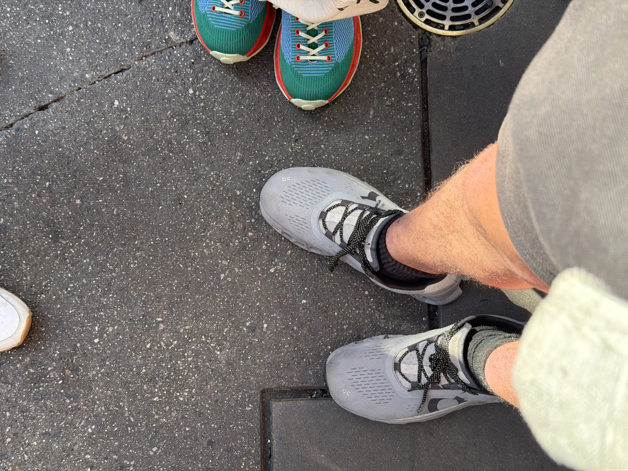 This image, taken in an outdoor setting likely within Anaheim, United States of America, focuses primarily on footwear. The composition is a close-up, high-angle shot of pavement with three distinct pairs of shoes visible.
The nearest pair of shoes are light gray running shoes with black and white patterned laces, worn by someone whose hairy legs are partially visible. The individual is also wearing gray shorts and a white cloth, possibly a towel or bag, is hanging down the side.
A second pair of shoes, positioned further away, are brightly colored running shoes featuring a combination of blue, green, and red hues with white laces. A third, single, white sneaker is partially visible in the lower left corner of the frame.
The ground appears to be a gray, asphalt-like surface with a scattering of small white specks. A metal grate is seen near the top right, adjacent to the colored sneakers, suggesting the location could be a street or sidewalk in an urban area. Overall, the lighting suggests it is daytime.