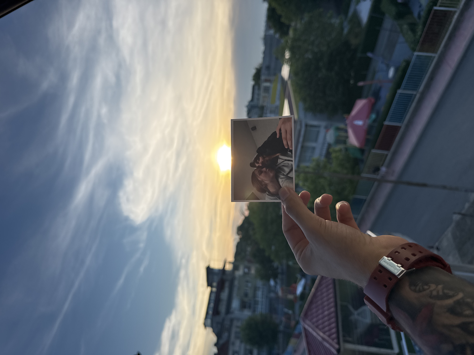 A hand holds a small, square photograph in front of a sunset sky over a city. The photograph shows a close-up of two people, appearing to be a couple, in an intimate embrace. The hand holding the photograph has a visible tattoo on the forearm and is wearing a maroon watch. The background of the main image is a blurred cityscape, with buildings and trees visible. The sunset is bright, casting a warm orange and yellow glow across the sky, with wispy clouds. The overall scene captures a personal moment superimposed against a wider urban landscape at dusk, with the sun positioned just above the horizon. The city of Tsarevo, Bulgaria, can be identified by the distinct architecture and coastal features visible in the blurred background, including red-tiled roofs and buildings with balconies, characteristic of the region.