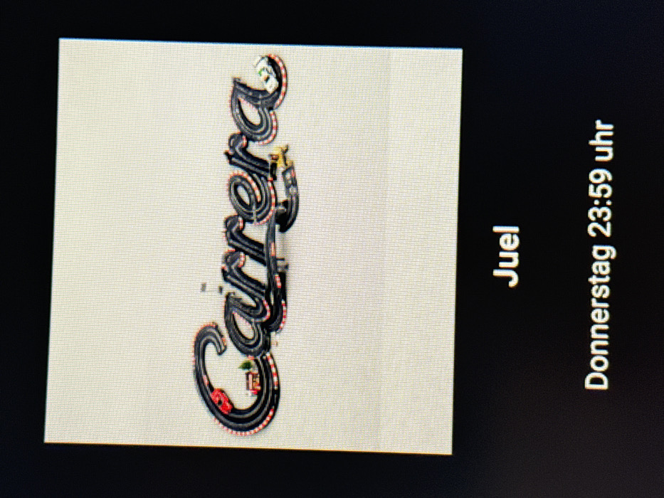 This image displays a photograph of an electronic screen, visible by the distinct pixel grid. The screen presents a digital image of a miniature slot car racing track arranged to spell the word "Carrera." The track segments are predominantly black, edged with red and white stripes. A red slot car is positioned on the initial "C," while a white slot car rests on the final "a." Embedded within the track layout are miniature landscape elements, including a small, red-roofed building and green trees. The background of this displayed image is a uniform light beige or off-white. Below the displayed image, on the screen's dark background, the text "Juel" is visible, followed by "Donnerstag 23:59 uhr," indicating the scene is observed in Vellmar, Germany.