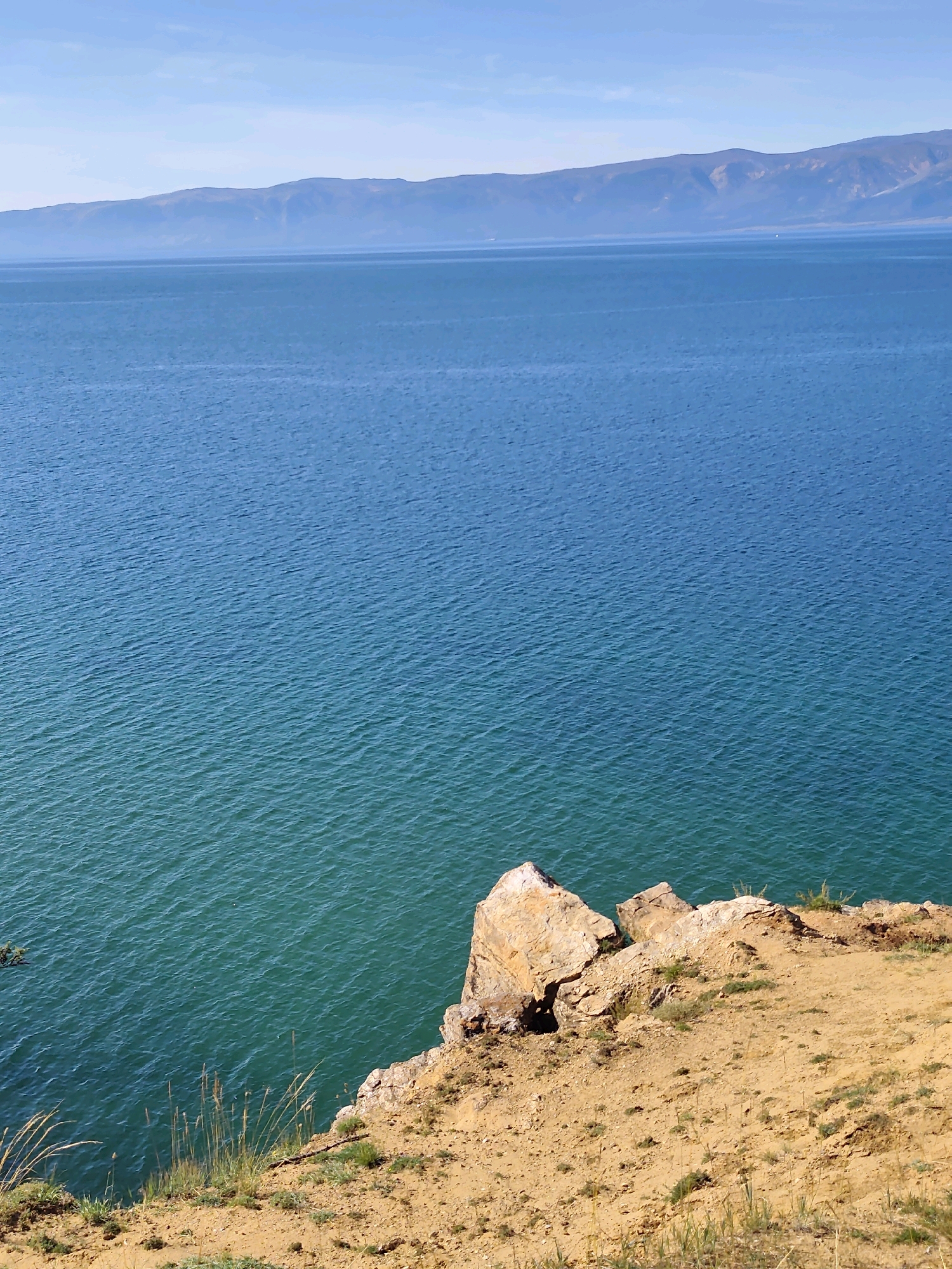 This image depicts a serene landscape with a body of water, likely Lake Baikal, meeting a rugged coastline. The water is a deep blue, transitioning to turquoise closer to the shore, with gentle ripples indicating calm conditions. In the foreground, a sandy cliff face rises from the water, adorned with sparse dry grass and a cluster of large, weathered rocks. The cliff is a warm, sandy brown color, with visible layers and textures suggesting erosion. Across the vast expanse of water, a hazy mountain range forms the horizon, its peaks softened by atmospheric perspective under a clear, pale blue sky. The scene is captured from a high vantage point, looking down and across the water towards the distant mountains, giving a sense of scale and openness. The city of Kharantsy, located on Olkhon Island in Lake Baikal, is known for its dramatic landscapes and this image appears to be a representative view of its natural beauty. There are no people or animals visible in the image.
