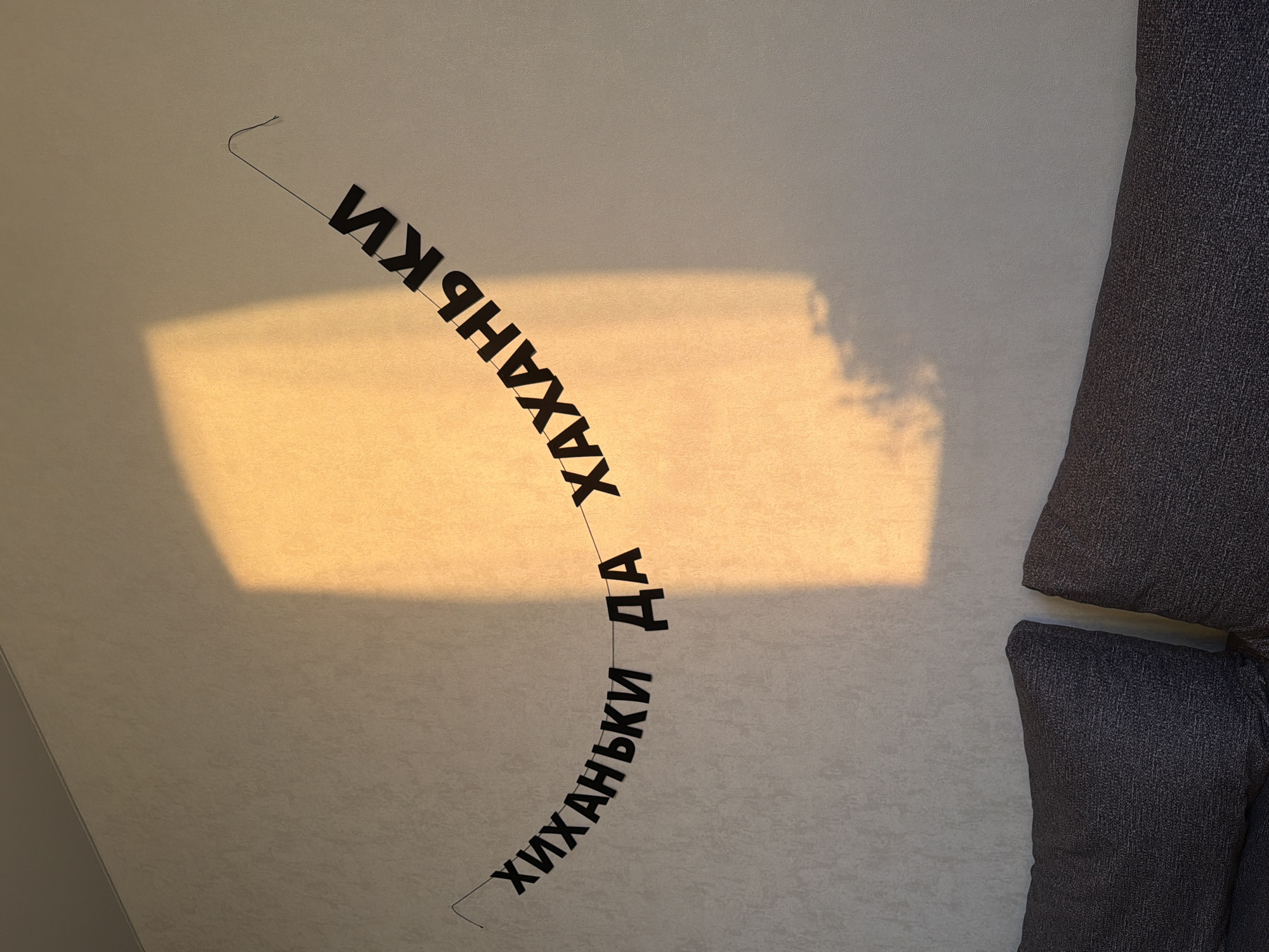 A view of a wall with a light brown shade on the left side. The word "хиханьки да хаханьки" is printed on the wall in black Cyrillic letters, strung on a black thread. The right edge of the image shows a gray cushion. The location is in an unknown city, and no further details are available.