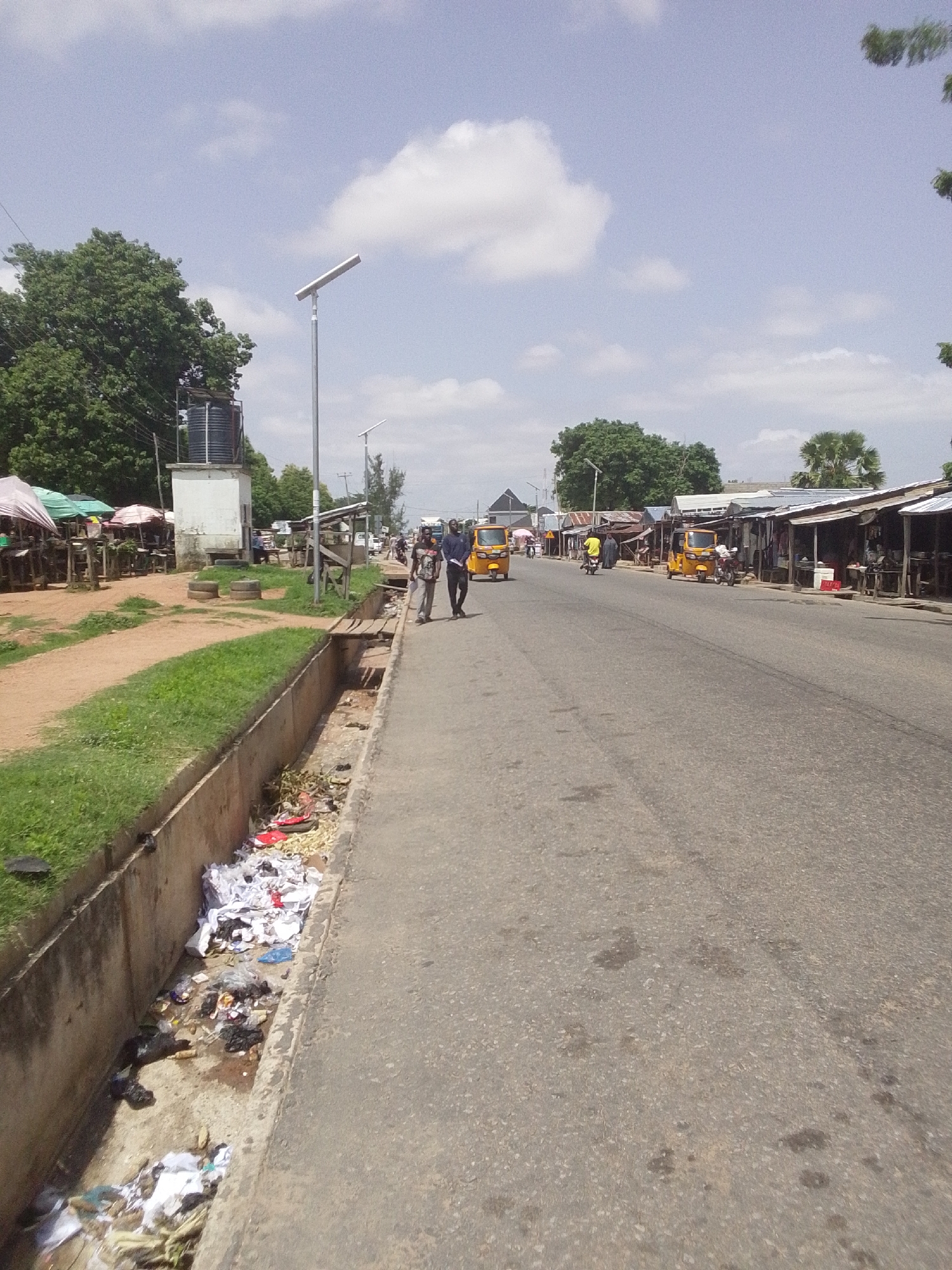 The image shows a street scene in Jalingo, Nigeria. On the left side of the road, there is a market with stalls and umbrellas, and a large water tank on a concrete structure. Adjacent to the market is a dirt path with grass bordering a concrete drainage ditch filled with debris and trash. On the right side of the road, a paved street runs into the distance, lined with shops. Several yellow auto-rickshaws, also known as *keke napeps*, are visible on the street, along with two men walking on the sidewalk. A solar-powered street light is present near the market area. The sky is partly cloudy with scattered white clouds.