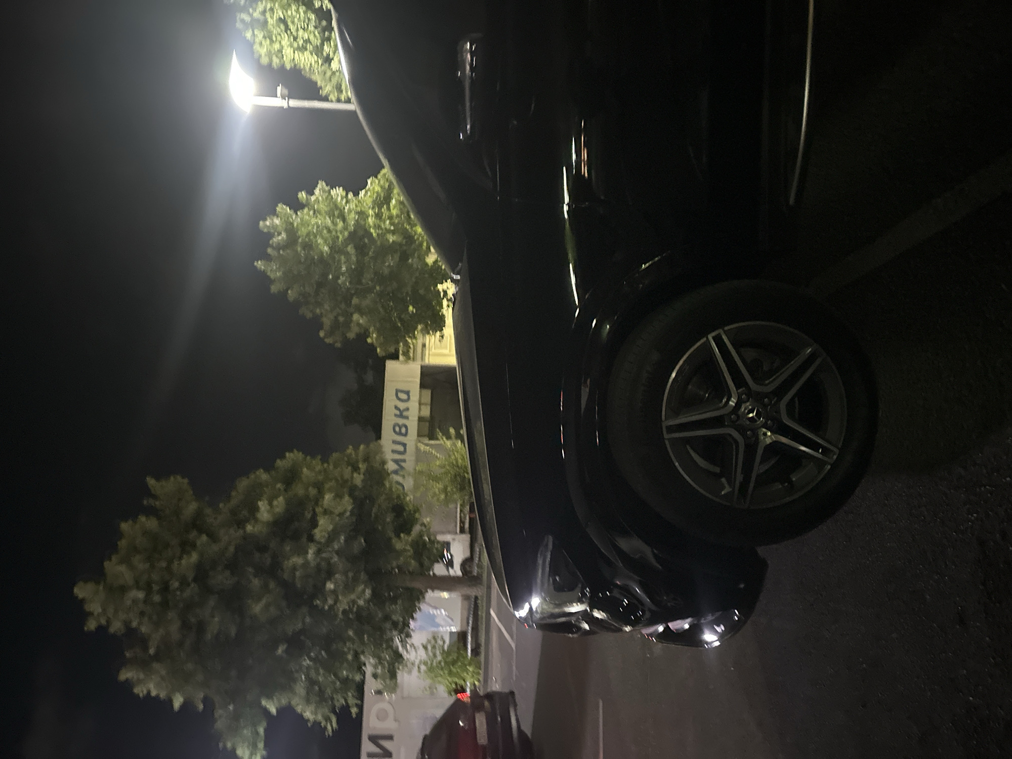 The image depicts a black Mercedes-Benz GLE parked at night in a dimly lit outdoor area, possibly a parking lot or a street. In the background, there is a building with signage that appears to be in Cyrillic script, with the word "МОЙКА" (MOIKA), meaning "car wash" or "washing," visible on a white facade. Several large trees with green foliage are present, illuminated by artificial lighting. A street lamp is visible in the upper right portion of the image. The ground surface is asphalt. Another dark-colored vehicle is parked to the left of the Mercedes. The overall scene suggests an urban environment in Ruse, Bulgaria, at night.