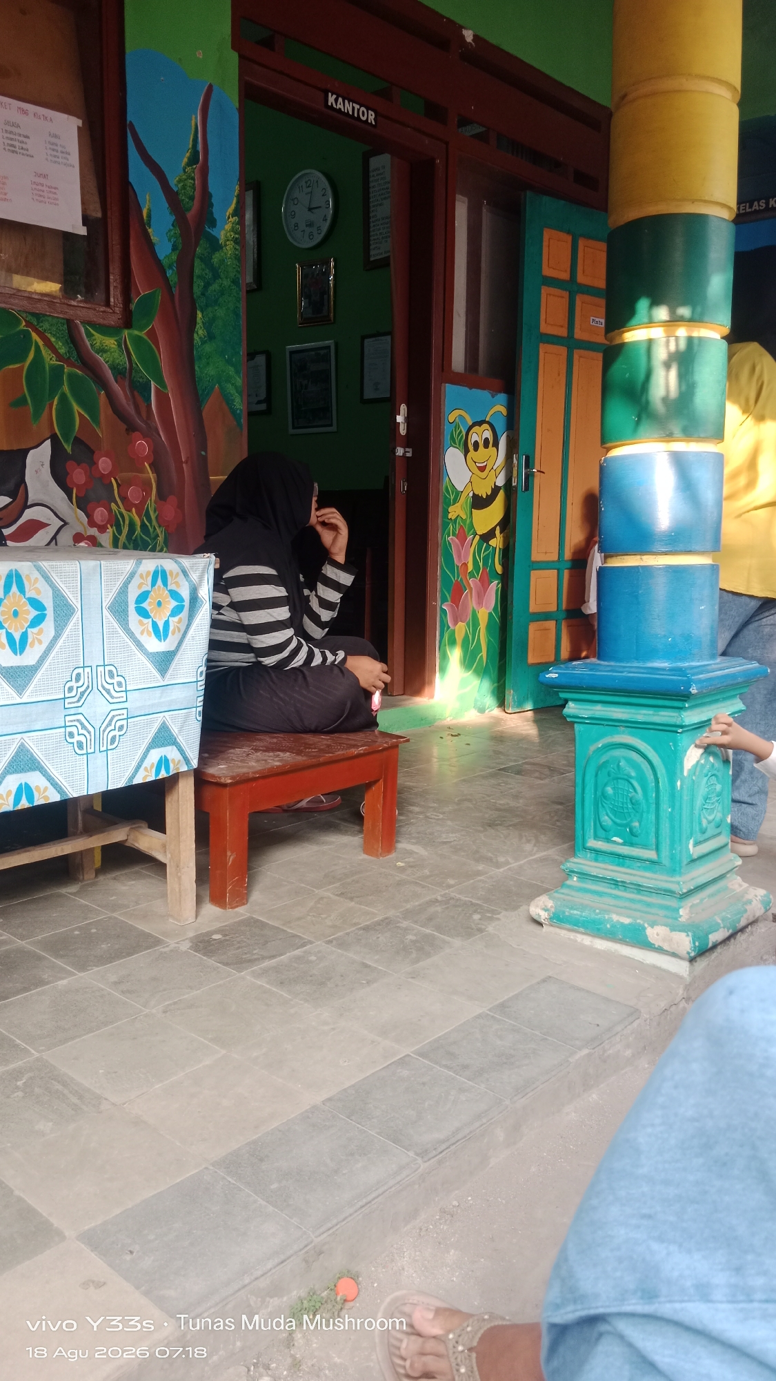 The image depicts an outdoor scene in front of a building. To the left, a woman wearing a black hijab and a striped shirt is sitting on a low wooden bench. A table with a patterned cloth is in front of her. The building facade features colorful murals, including trees and flowers on the left, and a bee character with flowers on the right, near a doorway labeled "KANTOR" (Office).
To the right of the doorway, a multi-colored pillar made of stacked circular objects (yellow, green, and blue) stands. A small green decorative base is at the bottom of the pillar. The ground is paved with square tiles. In the background, through an open doorway, a clock is visible on the wall of the office.
The bottom of the image shows a timestamp "18 Agu 2026 07.18" and text "vivo Y33s, Tunas Muda Mushroom," suggesting the photograph was taken at this time and location, possibly in a school or community setting in Tanjung, Indonesia. The presence of a child's hand reaching towards the pillar in the lower right corner indicates another person is present, out of frame. The overall atmosphere suggests a bright, possibly sunny day.