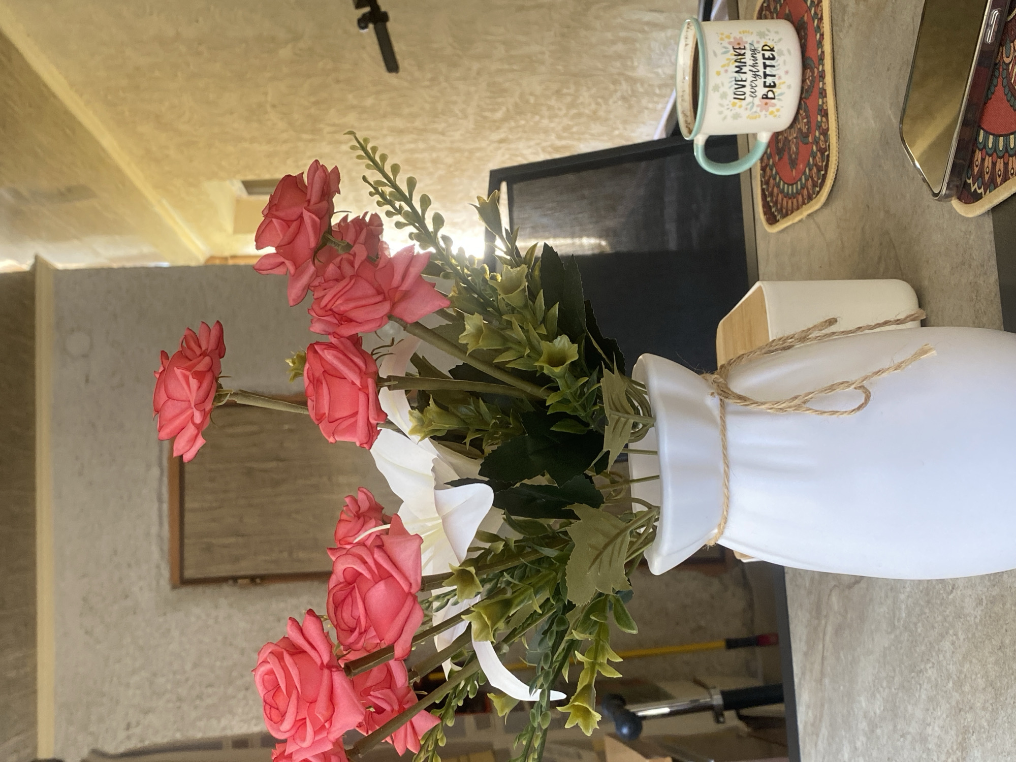 A still life scene unfolds on a textured, grey table, likely outdoors given the natural light and background elements. In the foreground, a white ceramic vase, adorned with a simple twine tie, holds a vibrant bouquet of artificial pink roses and white lilies, interspersed with green foliage. To the right of the vase, a white rectangular object, possibly a diffuser or coaster, is partially visible. Further to the right, a mug with a teal rim sits on a circular, patterned coaster, containing dark coffee. The mug's inscription reads "LOVE MAKE EVERYTHING BETTER." Below this, another patterned coaster supports a smartphone, its reflective screen tilted upwards. The background features a textured, light-colored wall with a wooden door inset, and a glimpse of a window with bright light streaming through, suggesting a building's exterior or an open-air space. A black object, potentially a part of a stand or easel, is visible behind the flowers. The overall impression is one of a serene, aesthetically pleasing arrangement, likely set in a garden or patio area in Lagodekhi, Georgia.