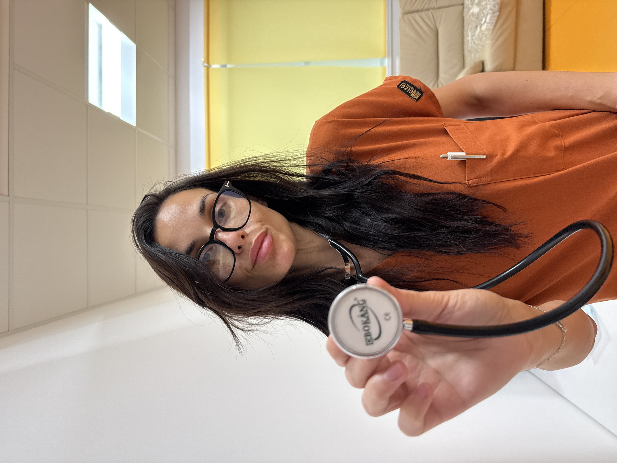 The image shows a woman in a professional setting, likely in Pleven, Bulgaria, looking directly at the viewer with a neutral to slightly smiling expression. She has long, dark hair and wears black-framed glasses. She is dressed in an orange scrub top or uniform shirt. A patch on her left sleeve displays the text "GIZGI," and a silver pen, which appears to be Montblanc, is tucked into her chest pocket.
She extends her right hand towards the camera, holding a stethoscope. The silver-toned bell of the stethoscope features the brand name "EBOKANG" and the "CE" mark.
The background reveals an indoor environment with light-colored walls, possibly tiled, and a bright rectangular light fixture on the upper left. To the right, there's a glimpse of yellow blinds or a yellow wall, and a section of soft, upholstered furniture in a light color. The lighting is bright and even, suggesting an indoor space during the daytime. The perspective of the image is rotated. The scene conveys a professional and potentially medical context.