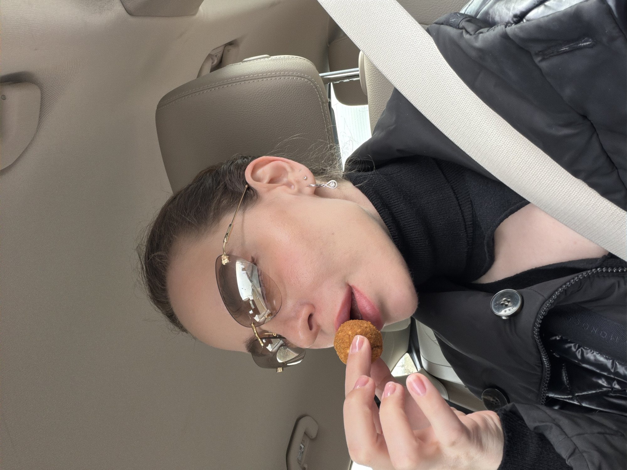 A woman is inside a vehicle, wearing sunglasses and a black puffer jacket with a black turtleneck sweater underneath. She is wearing a seatbelt. She holds a small, round, brown food item between her fingers and her mouth is open as if to eat it. The interior of the car is beige. No location in Zhedochi, Russia, is discernable.