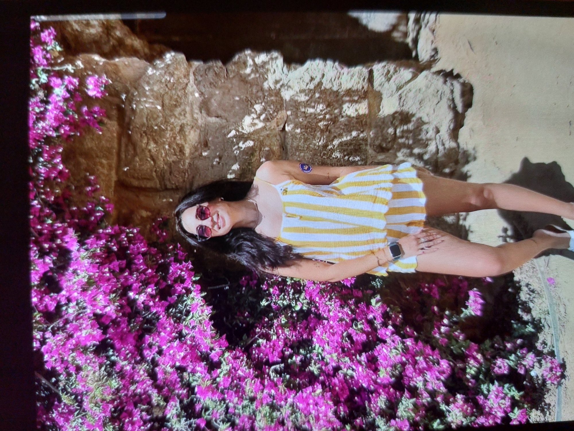A vibrant outdoor portrait shows a smiling young woman posing amidst colorful bougainvillea flowers and a textured rock formation, likely in Tsarevo, Bulgaria. The woman, with long dark hair, is wearing a yellow and white striped sundress or romper, red-tinted sunglasses, and a dark smartwatch on her left wrist. She is looking directly at the camera with a happy expression.
To her left, a dense cluster of bright pink/purple bougainvillea flowers covers a significant portion of the scene, adding a pop of color. Behind her and to her right, a rugged, light-brown rock wall or cliff face provides a natural backdrop. The ground appears sandy or dirt-covered.
The scene is brightly lit by what appears to be direct sunlight, indicated by distinct shadows cast on the ground and rock wall, suggesting it's daytime. A small blue circular sticker or patch with a white design is visible on her left upper arm. The overall atmosphere is cheerful and relaxed, capturing a moment in a beautiful, sunny outdoor setting.