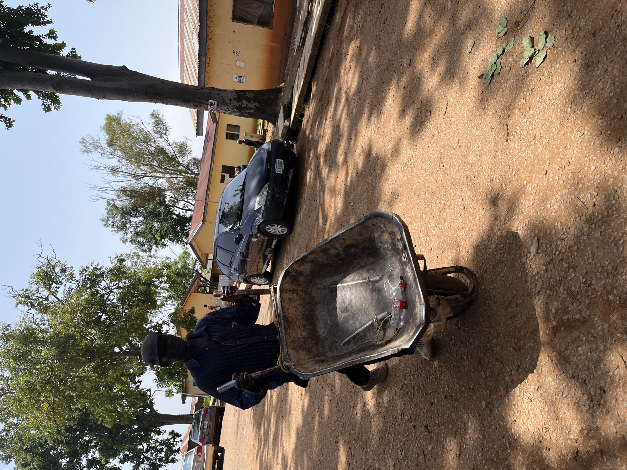 A man wearing a dark striped shirt, dark pants, and a cap pushes a wheelbarrow filled with two plastic bottles. He is walking on a dirt road in front of yellow buildings with corrugated metal roofs. A dark blue Ford Focus is parked on the road to his right, and other cars are visible in the background. Trees and sunlight create shadows on the ground. The scene appears to be in Jos, Nigeria.