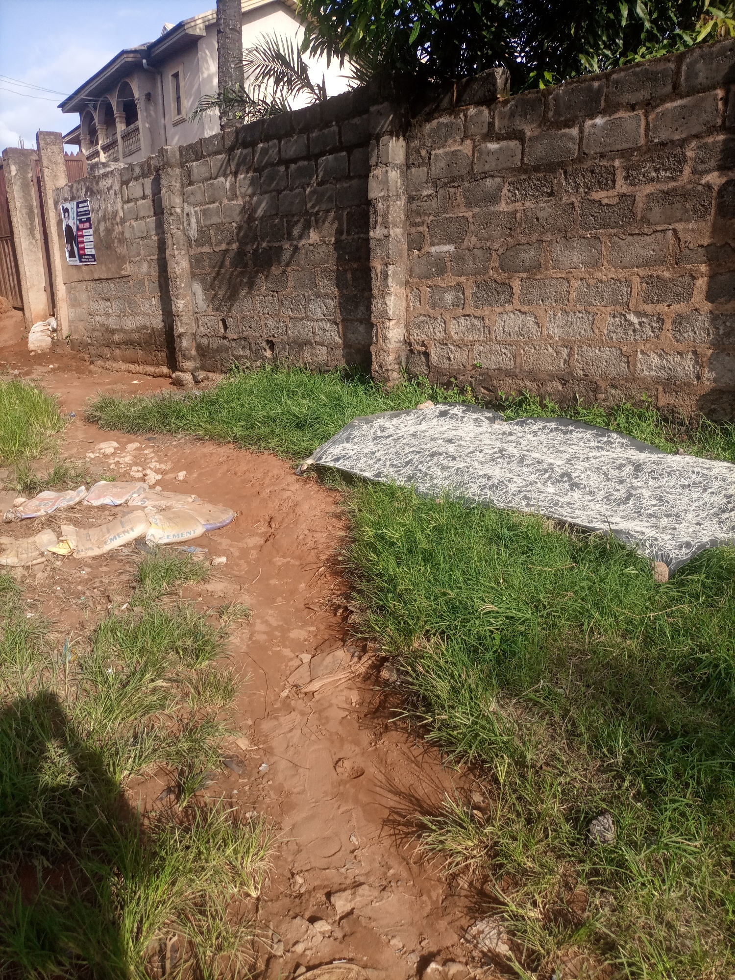 The image depicts an outdoor scene in Nkpor, Nigeria, characterized by a dirt path, overgrown grass, and a weathered brick wall. The wall appears to be part of a residential property, with glimpses of a building and a fence visible to the left. On the wall, a poster with text and images is affixed, suggesting a public announcement or advertisement. In the foreground, several torn cement bags lie on the ground, indicating construction or waste materials. Further into the scene, a large piece of dark fabric, possibly a tarp or net, is spread out on the grass, covered with a dense layer of white, stringy material, which appears to be drying in the sun. The overall environment suggests a somewhat unkempt or utilitarian area within the urban landscape.