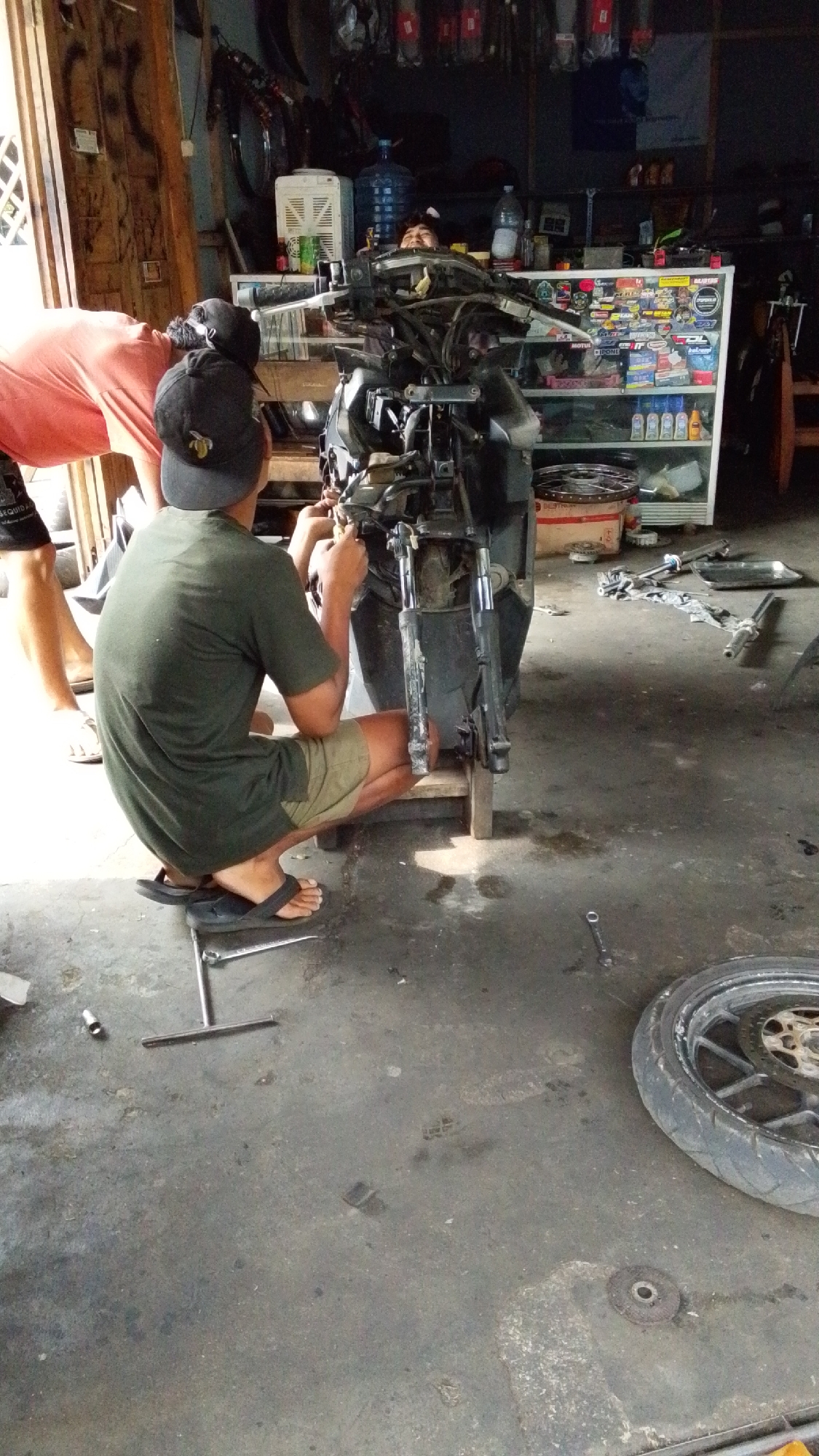 In an open-air workshop or garage in Cintaratu, Indonesia, a person wearing a green t-shirt and olive shorts crouches down, working on the front end of a disassembled motorcycle. They are holding a tool in their right hand, actively engaged in repairing the vehicle. Another person, partially visible and wearing an orange shirt and black shorts, stands behind the motorcycle, also appearing to be involved in the repair process. The motorcycle's front suspension and handlebars are exposed, indicating mechanical work is underway. Shelves filled with various items, including bottles, are visible in the background, along with large water jugs and a white appliance, likely an air cooler. The floor is concrete, with tools, including a wrench, scattered around. A motorcycle wheel lies on the ground to the right of the scene. The overall environment suggests a functional, working garage space.
