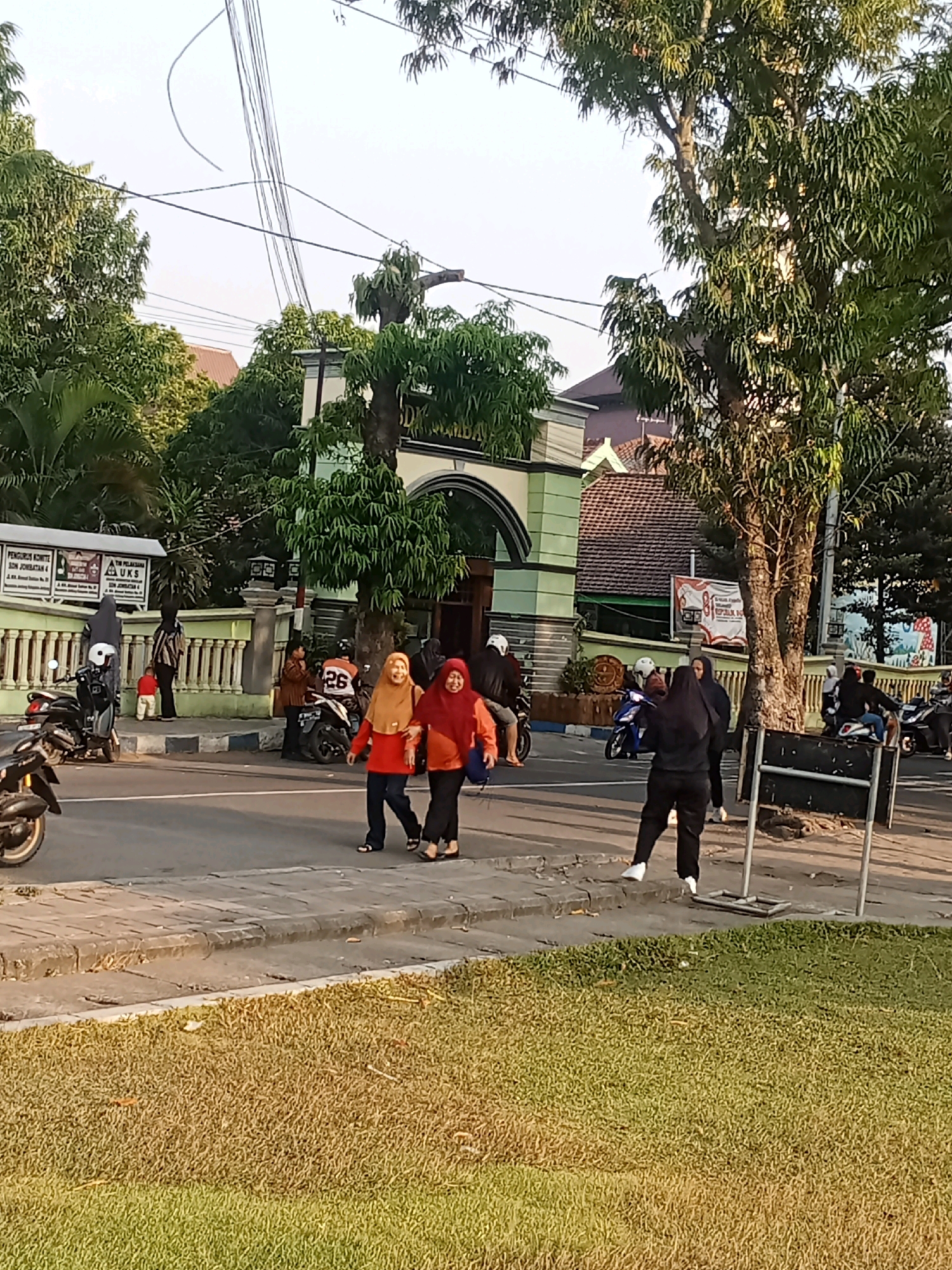 The image depicts a street scene in Kaliwungu, Indonesia, with a building featuring an archway entrance and a green facade. Several individuals are present, some on foot and others on motorcycles. In the foreground, two women wearing headscarves are walking, with one smiling and holding the other's arm. Two motorcycles are parked on the left side of the road, and more motorcycles are visible further down the street. A patch of grass is in the lower part of the frame. Overhead power lines are visible crisscrossing the sky. The overall atmosphere suggests a typical day in a residential or public area.