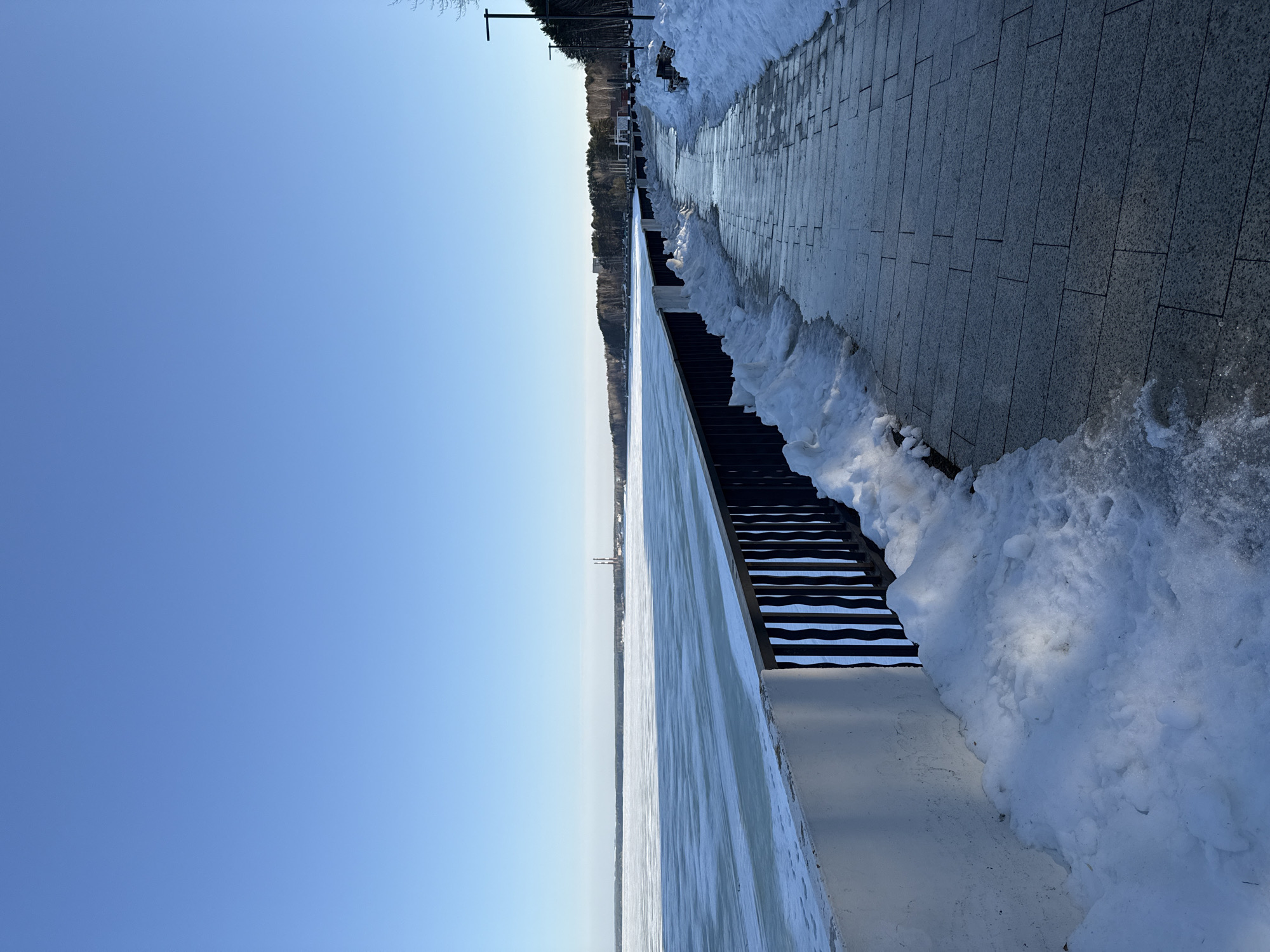 The image depicts a winter scene at the city of Chaikovsky, Russia. A paved walkway covered in snow runs along the right side of the image. Adjacent to the walkway is a metal and concrete retaining wall, partially covered in ice and snow. Beyond the wall, there is a frozen body of water, possibly a river or lake, stretching towards the horizon. The sky is a clear, light blue. A line of trees is visible in the distance.